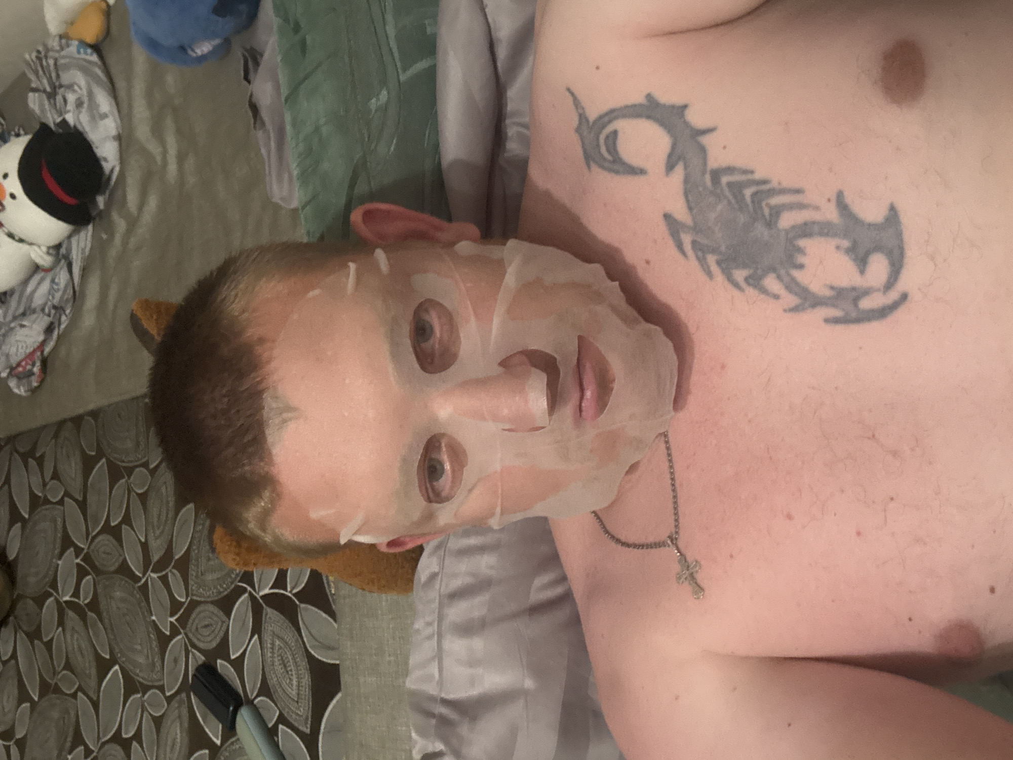 A man with light skin and short brown hair is lying down, facing the camera. He is wearing a white sheet face mask that covers most of his face, leaving his eyes, nose, and mouth visible. A silver cross necklace hangs around his neck. On his chest, a scorpion tattoo is visible on the right side. The background appears to be a bed with various pillows and stuffed animals, including a snowman and a blue plush toy. A patterned blanket or sheet is also visible in the background. The lighting suggests an indoor setting, likely a bedroom.