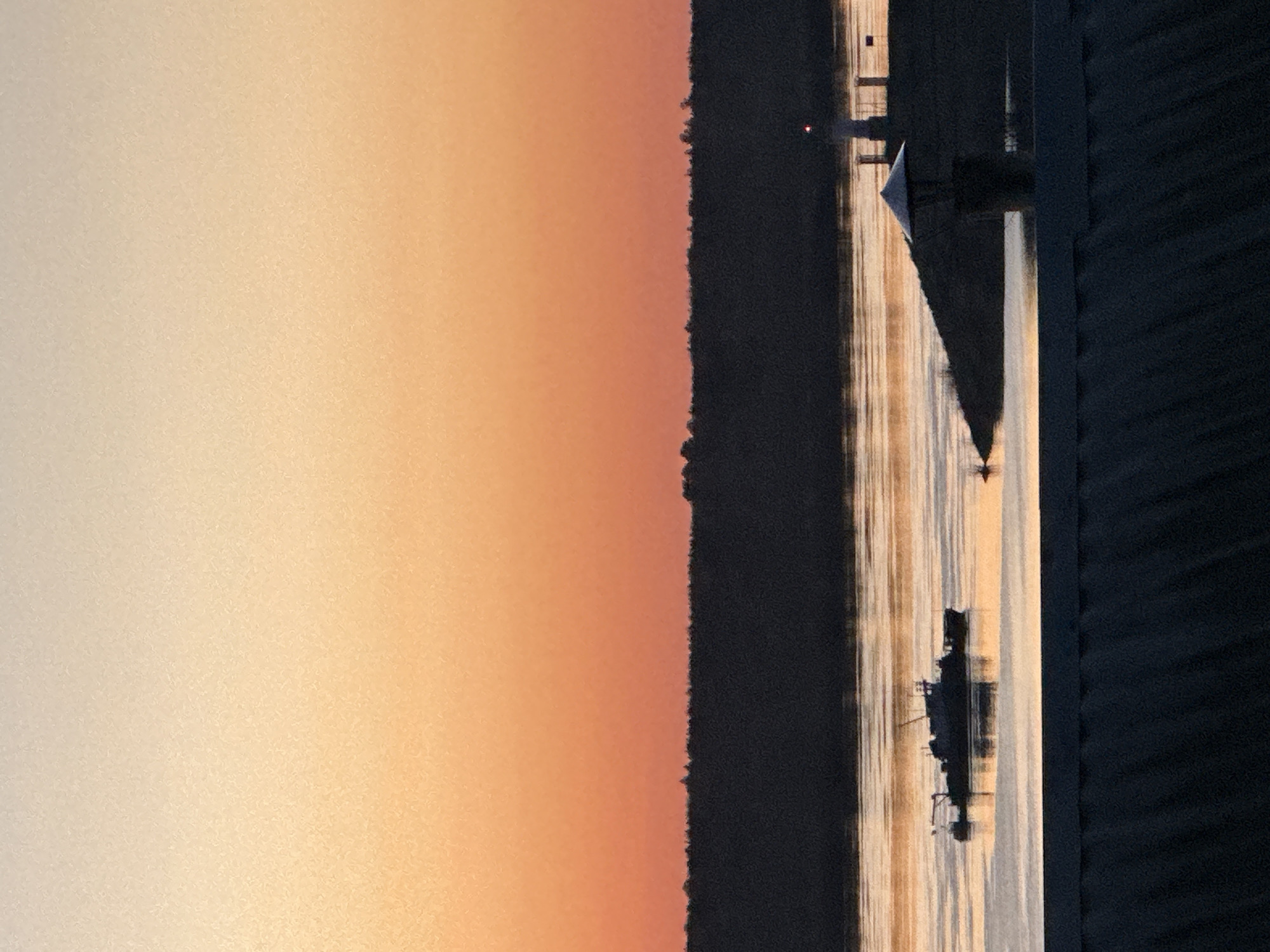 Here is a description of the image:
The image shows a body of water, likely a river or lake, reflecting the sky during sunset. The sky transitions from a light peach color at the horizon to a warm orange. A dark, silhouetted treeline is visible on the far bank of the water. In the water, there are reflections of what appears to be a building with a triangular roof and some smaller structures. There is also what looks like a small ship or boat. A dark area on the right could be a road or bank. The overall scene suggests a serene, rural landscape at sunset, potentially situated near the city of Chaikovsky, Russia.