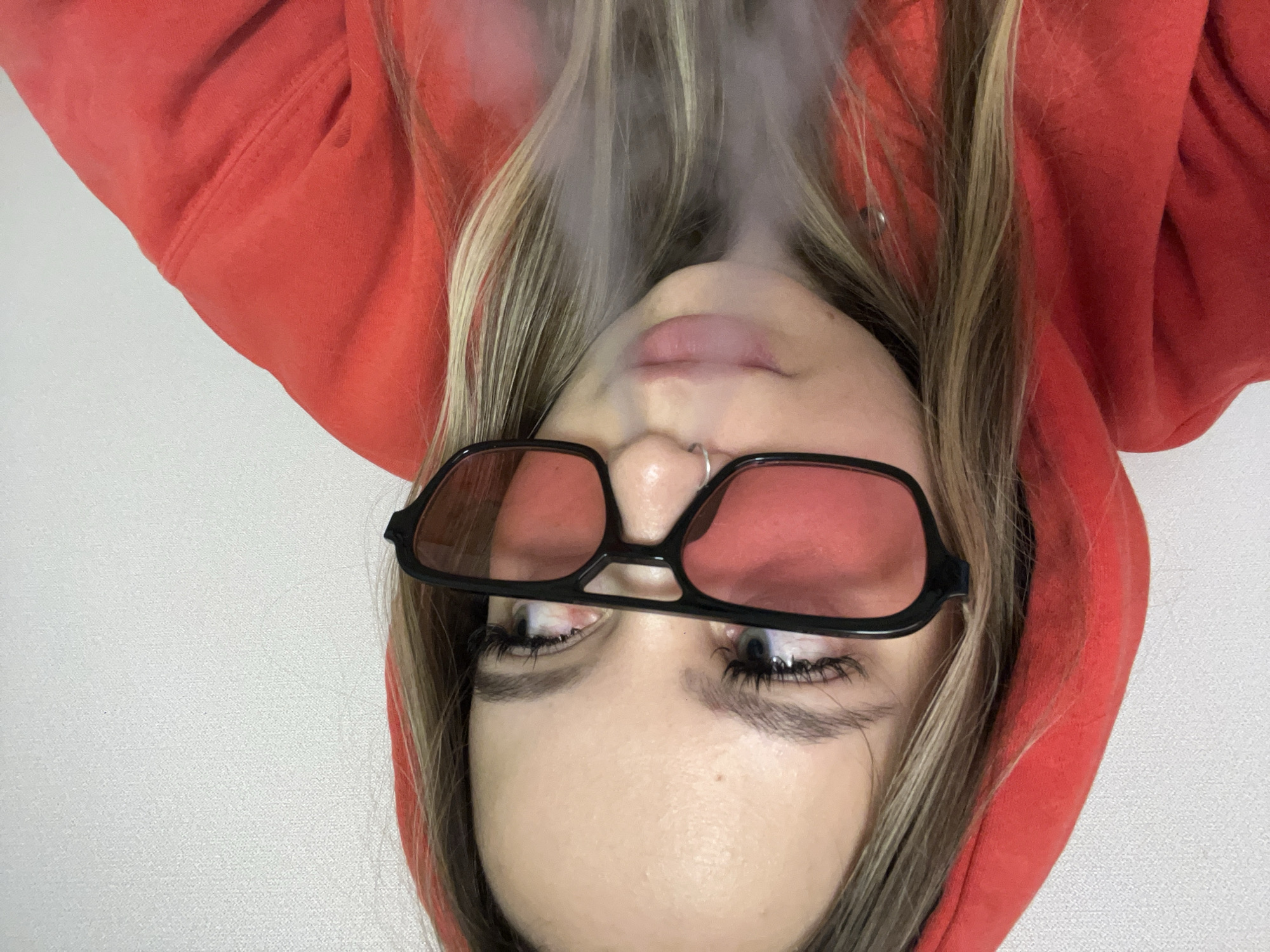An inverted close-up portrait features a young person, likely female, with long blonde and light brown hair. The subject is wearing a vibrant red hooded sweatshirt, with the hood framing their face. They are sporting black-framed sunglasses with distinct red-tinted lenses. A silver hoop nose ring is visible on their left nostril.
The central activity captured is a plume of white smoke or vapor exhaling from the person's mouth, rising upwards towards the top of the frame and partially obscuring their lower face. Their light-colored eyes, visible beneath the sunglasses, appear to be looking to the side. Notable details include visible eyelashes, defined eyebrows, and full lips partially hidden by the smoke.
The background is a plain, light-colored wall, suggesting an indoor setting. There are no specific location cues within the image itself, but the context provided places the scene in Krasnodar, Russia. No text is visible in the media file. The overall impression is one of a casual and stylistic moment.