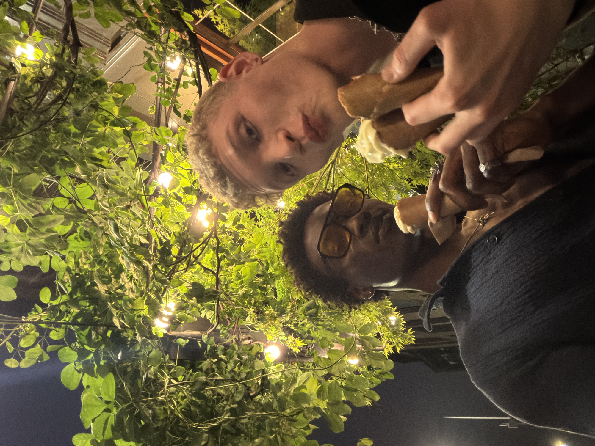 An image, rotated 90 degrees counter-clockwise, depicts two individuals from a low-angle perspective, possibly a selfie taken from below, looking up towards the camera. The scene is set at night, illuminated by numerous string lights nestled amongst abundant green foliage, suggesting an outdoor patio or garden area in London, United Kingdom.
On the left side of the frame (when viewed upright), a person with light, curly hair and fair skin is prominently featured, making a playful pouting expression directly at the camera. They wear a dark top, with a subtle chain visible around their neck. This individual holds a cream-filled pastry, likely a cannoli, in their right hand, bringing it towards their mouth.
To the right, partially obscured by the first person's arm and the surrounding leaves, is another individual with darker, curly hair. They are wearing amber-tinted glasses and a dark, possibly ribbed, garment. This person also holds a similar cream-filled pastry. A ring is visible on one of their fingers as they hold the dessert.
The activity clearly shows both individuals enjoying pastries, captured in a candid and possibly playful moment. The overhead canopy of lush green leaves and vines, intertwined with the warm glow of string lights, creates an intimate and pleasant nighttime atmosphere. No text is visible in the image.