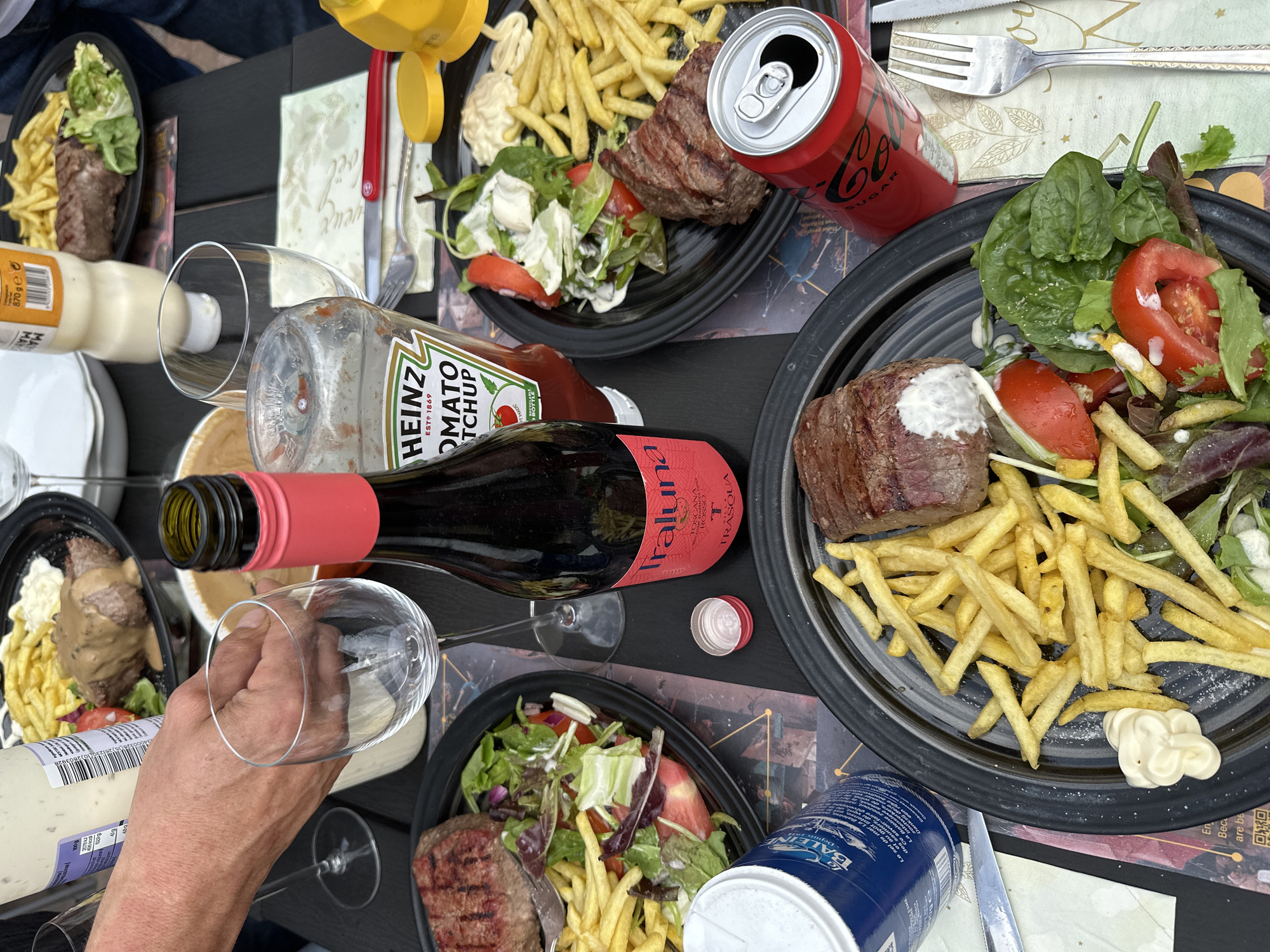 An outdoor table at a restaurant in Bruges, Belgium, is laden with plates of food and drinks. Two black plates contain grilled steak, french fries, and salad. A bottle of red wine stands between the plates. A ketchup bottle and a can of Coca-Cola are also visible. A hand is reaching for a wine glass. The setting suggests a casual meal, possibly lunch or dinner, enjoyed outdoors.