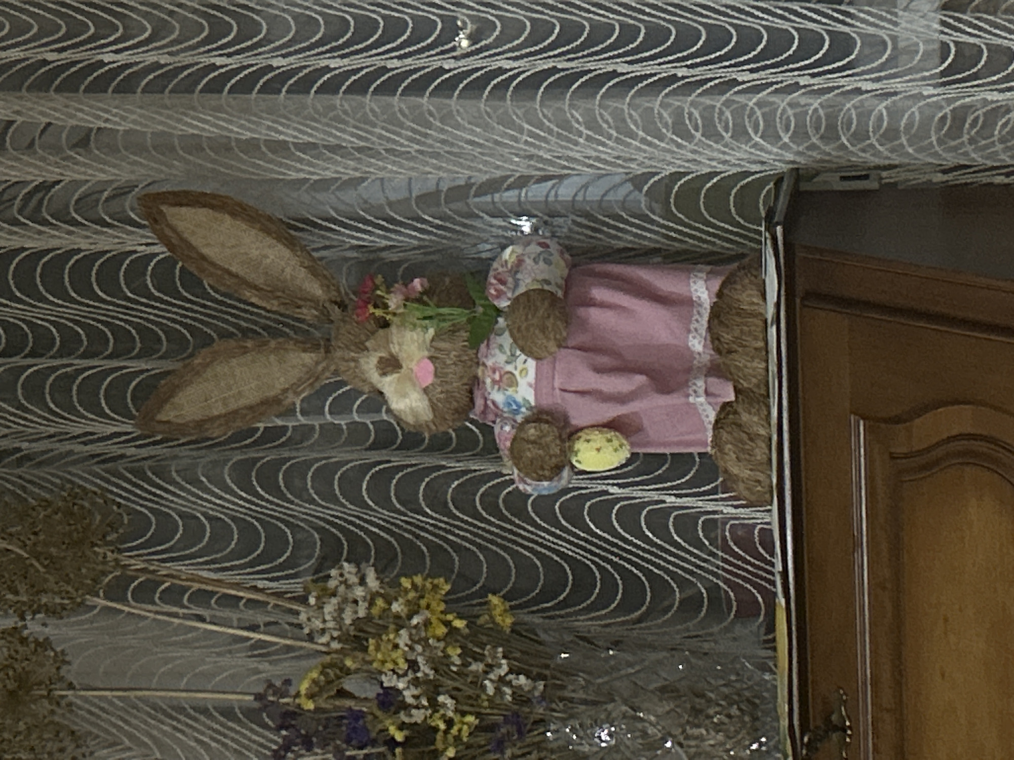 A decorative Easter bunny figure stands on a wooden surface, in front of a curtain with a wavy pattern. The bunny is brown with large ears, a pink nose, and a floral shirt with a pink dress. It holds a yellow Easter egg in its left hand and a small bouquet of flowers in its right. To the left of the bunny is a vase of dried flowers. The overall scene is indoors, possibly in a home in Răuțel, Moldova, judging by the mention of the city.