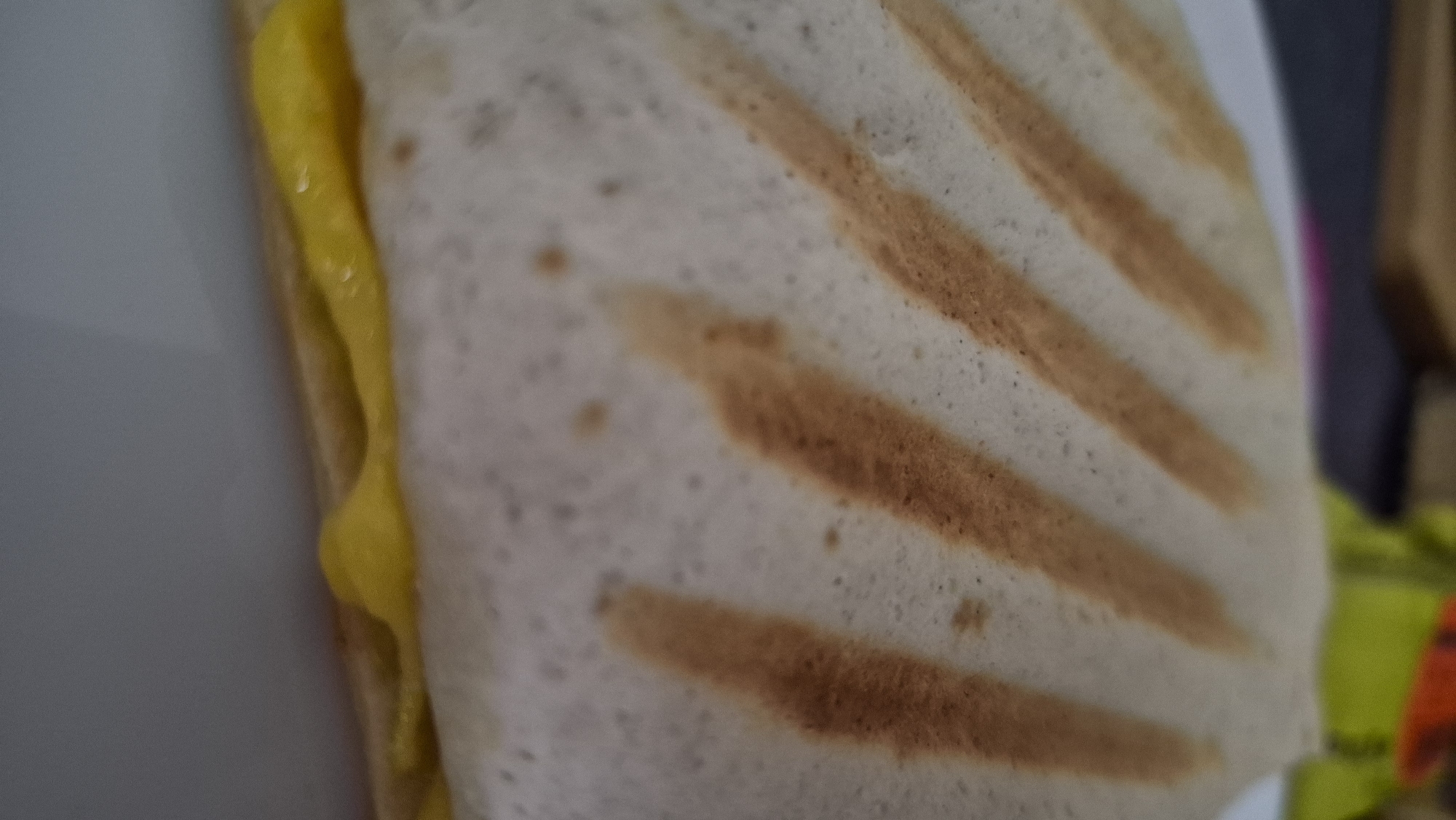 The image shows a close-up of a grilled tortilla wrap with yellow cheese oozing out from the bottom. The tortilla has grill marks running diagonally across the top. The wrap is placed on a white plate. The background is blurred, showing hints of a wooden surface and some colorful objects, possibly food packaging. There are no people or animals visible in the image. The location in Krauja, Latvia cannot be determined from the provided image.
