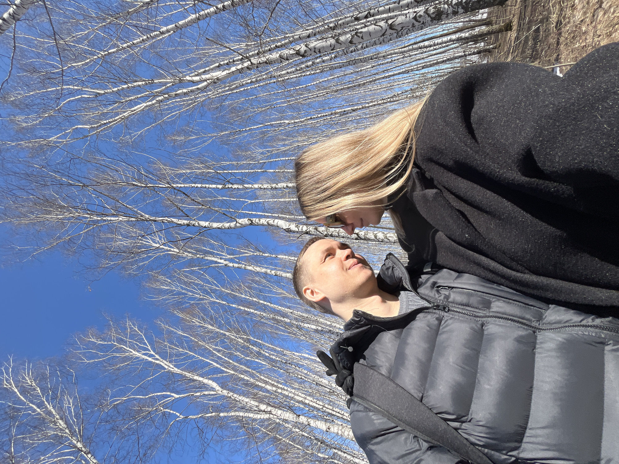 A man and a woman stand close to each other in a birch forest in Chaikovsky, Russia on a clear, sunny day. The woman is blonde and wears a dark coat. The man wears a gray puffer jacket and a dark shoulder bag. They are both gazing at each other. The forest consists of tall, thin birch trees with white bark, and the sky is a clear blue.