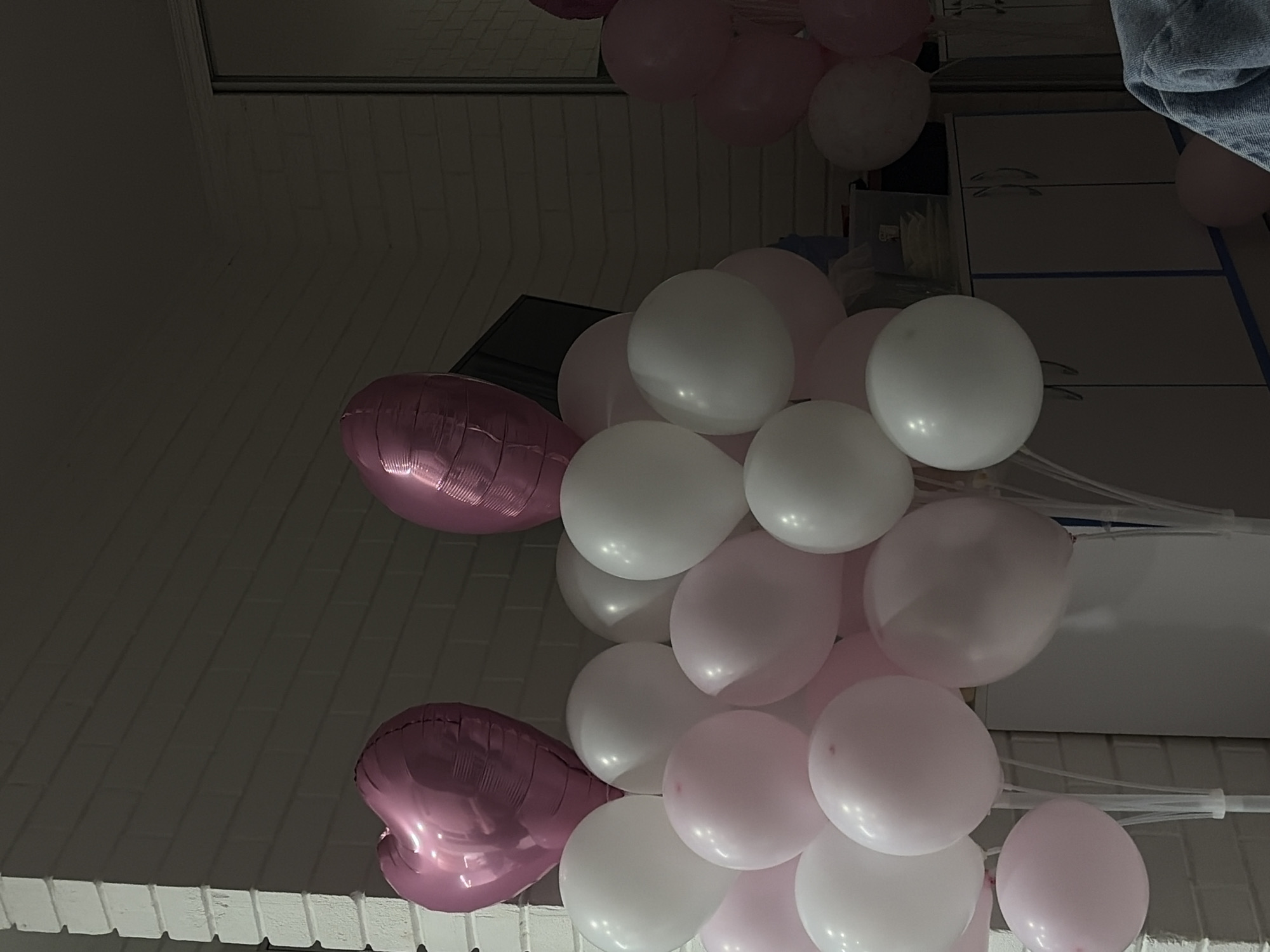 This is a close-up, medium shot of a balloon display with a brick wall in the background and a mirror reflecting a portion of the scene. The balloons are a mix of pink and white, with two prominent pink heart-shaped balloons at the top. The lower portion of the display consists of numerous round balloons, some white and some a lighter shade of pink, clustered together and attached to clear plastic stands. In the background, a mirrored closet door is visible on the right, reflecting some of the balloons and a dark cabinet. A person's shoulder and arm, wearing a denim jacket, are partially visible in the lower right corner of the frame. The lighting is dim, suggesting an indoor setting. The location is not specified in the image itself, and there is no indication of it being in Tiraspol City Council, Moldova.