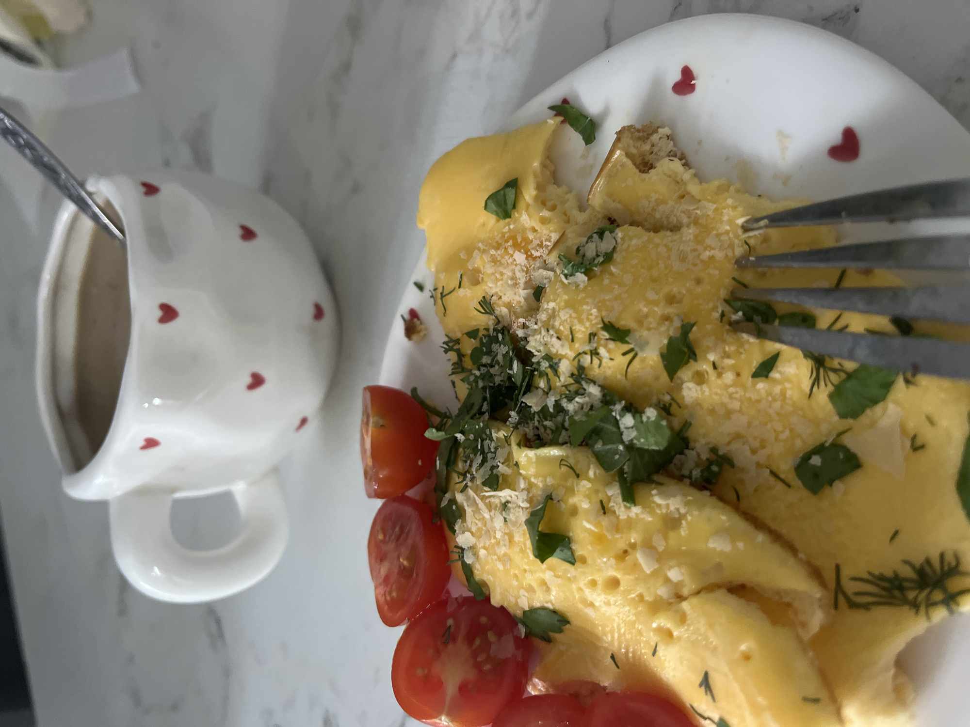 A still life captures a breakfast scene featuring a white plate with red heart designs holding an omelet garnished with chopped tomatoes, parsley, and grated cheese. A fork is inserted into the omelet. Adjacent to the plate is a white mug adorned with red hearts, containing a beverage with a spoon inside. The background is a textured white surface. The location is unknown, therefore, I cannot provide information about the city of Chaikovsky, Russia.