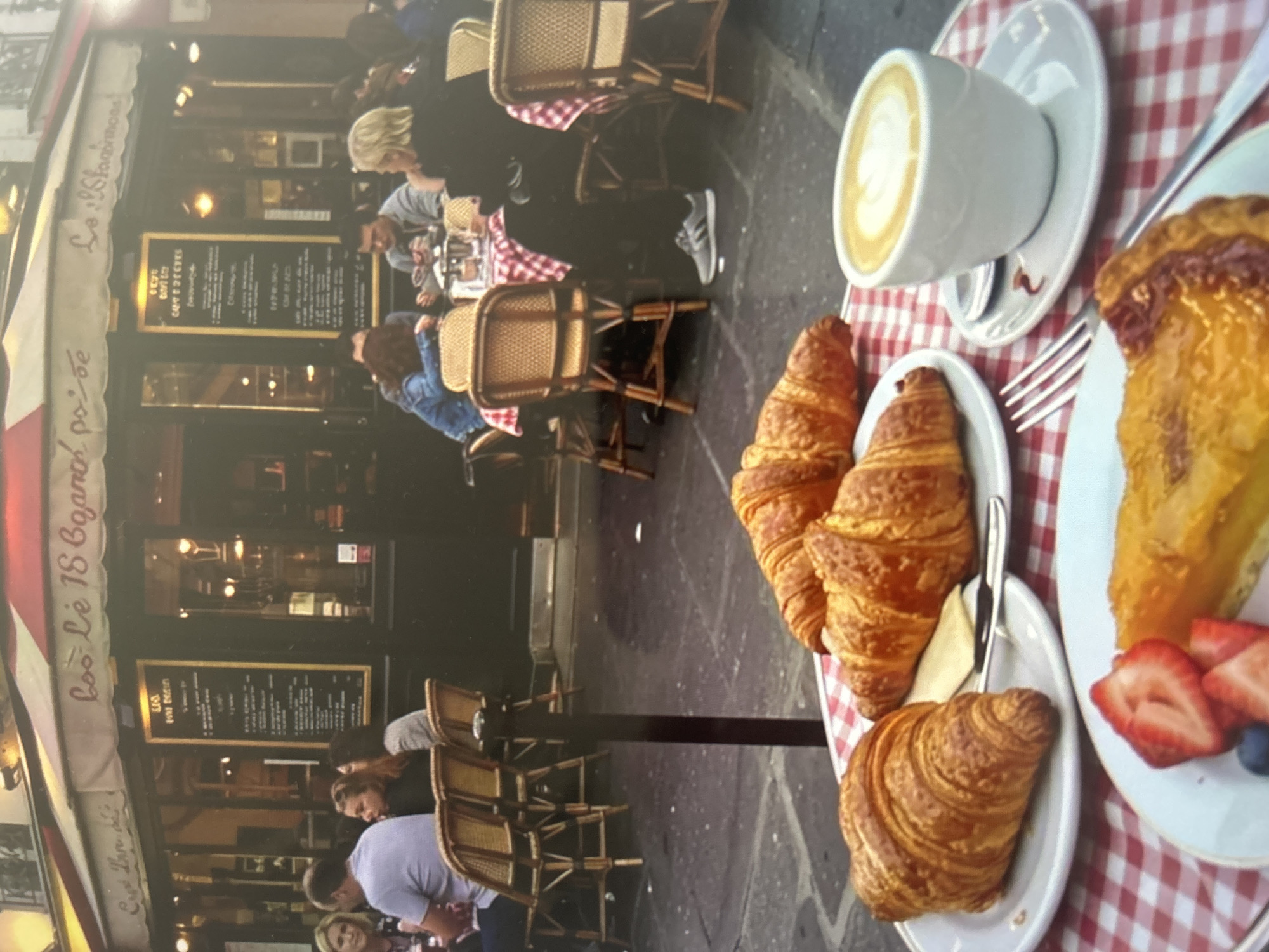 The image depicts an inviting outdoor cafe scene, likely in a European city, centered around a table set for breakfast or brunch. In the foreground, a red and white checkered tablecloth covers a small round table laden with delicious food. Three golden-brown croissants are arranged on white plates, one accompanied by a small pat of butter. Adjacent to them is a slice of what appears to be a fruit tart, possibly apple or pear, garnished with fresh red strawberries and blue blueberries, served on a white plate with a silver fork. A white ceramic cup filled with coffee, topped with delicate latte art, sits on its saucer.
In the midground, several individuals are seated at similar small tables with wicker-style chairs, enjoying the cafe's ambiance. They are engaged in conversation or observing their surroundings. One person's foot, wearing a grey Adidas sneaker, is visible under a table. The atmosphere appears relaxed and casual, suggesting a pleasant daytime gathering.
The background reveals the facade of the cafe itself, characterized by dark-framed windows and a traditional awning. A prominent black menu board with gold trim is displayed outside the cafe, listing various items and their prices, though the specifics are largely unreadable. Visible text on the awning clearly states "Le 18 Bogard," with "Café" and "Restaurant" also discernible beneath it. The overall setting suggests a pleasant morning or afternoon, with ample light for outdoor dining.