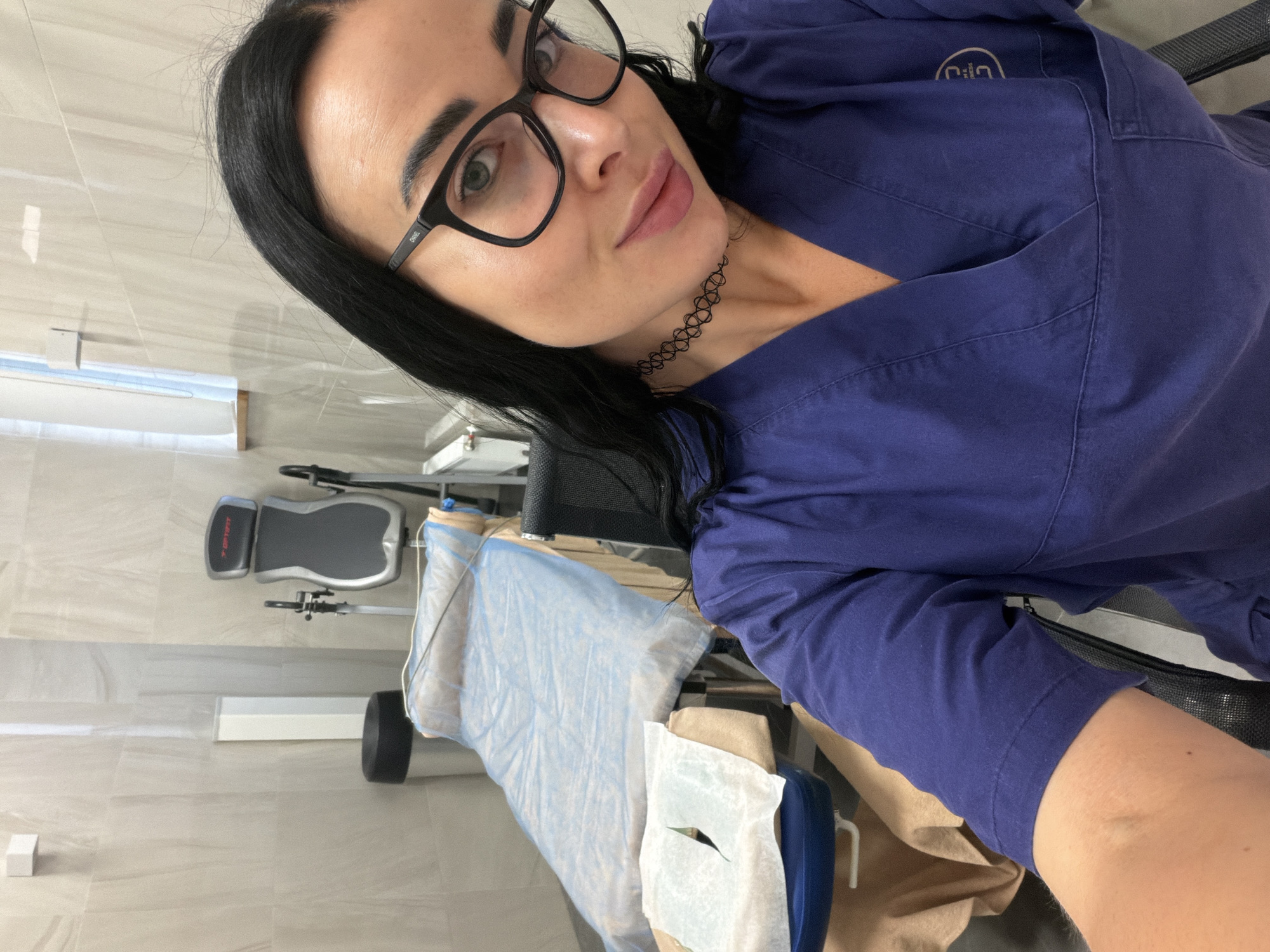 The photograph appears to be a selfie taken by a woman indoors. She has dark hair, wears glasses, and is wearing a blue scrub top. In the background, there is medical equipment, including an examination table covered with a blue drape and paper. The location is likely a medical examination room. The absence of context in the provided image prevents an accurate identification of the location within Saint Petersburg.