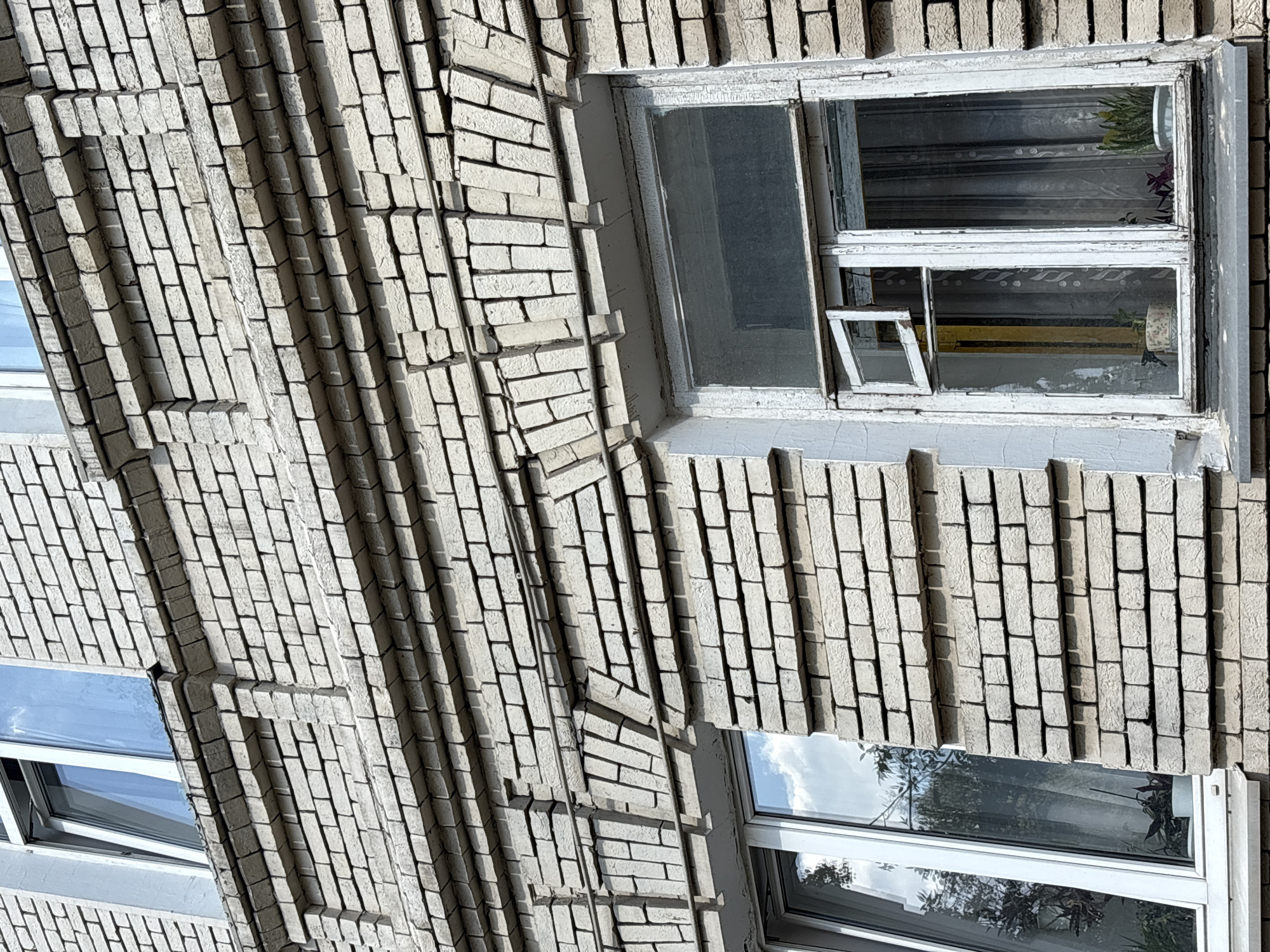 The image shows the facade of a brick building, likely an apartment building, in Moscow, Russia. The bricks are light in color and show some signs of weathering. The building features decorative brickwork above the windows, including horizontal bands and arched lintels. There are several windows visible; one is a modern white-framed window with a clear pane reflecting the sky and trees. Another is an older, white-painted wooden window with multiple panes, one of which is open, revealing curtains and houseplants inside. The scene is devoid of people or any activity.