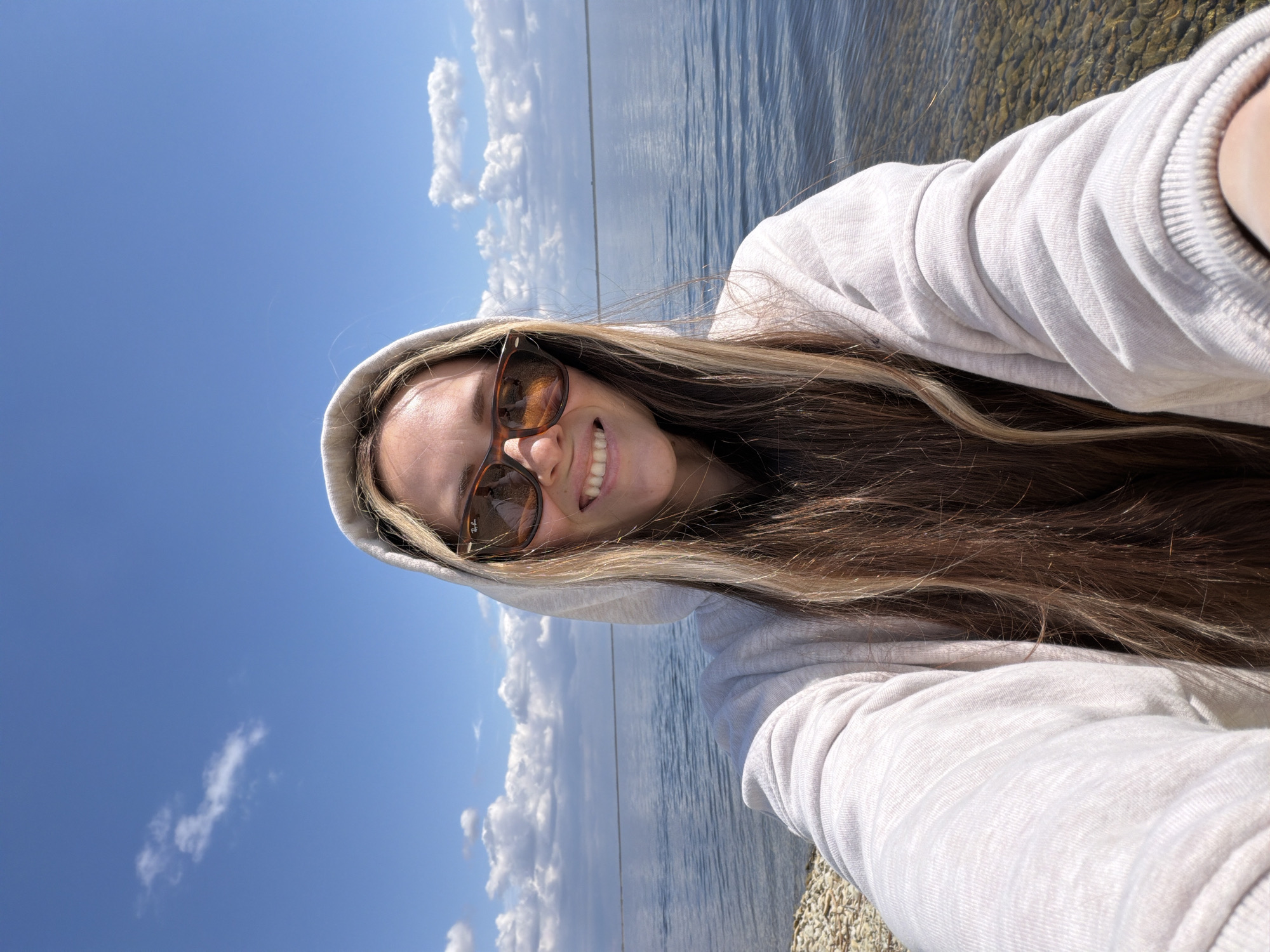 A woman wearing sunglasses and a hooded sweatshirt smiles for a selfie. The background shows a body of water, presumably the Volga River, as Kamyshin is situated on its banks in Russia. The sky is blue with scattered white clouds. Gravel or small rocks are visible at the bottom right edge, indicating a shoreline.
