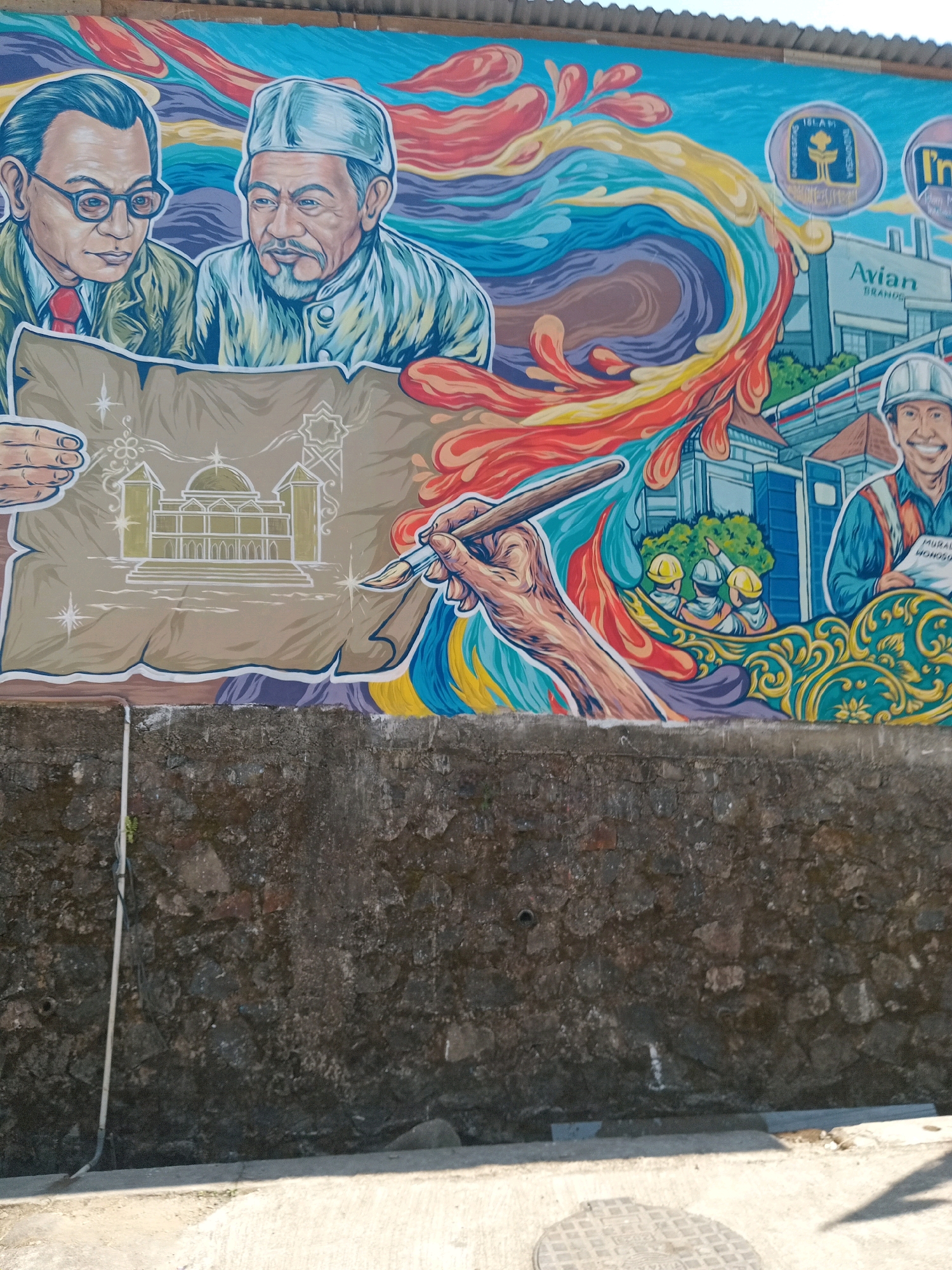 The image is a photograph of a vibrant mural painted on a wall in Maduretno, Indonesia. The mural depicts two distinguished men, likely historical figures or community leaders, poring over a blueprint or architectural drawing of a mosque. One man wears glasses and a suit, while the other wears a traditional cap. A hand holding a paintbrush is shown in the process of adding details to the drawing, suggesting a focus on planning and development.
The background of the mural is a dynamic swirl of colorful abstract shapes in shades of blue, red, yellow, and orange, representing energy and progress. Interspersed within the mural are various emblems and logos, including a circular emblem with text that appears to say "UNIVERSITAS ISLAM INDONESIA" and another circular logo with the "Avian" brand name, suggesting local institutions and businesses are recognized or involved. Several figures wearing hard hats and construction vests are depicted in the mural, pointing towards buildings, further emphasizing themes of construction and community building. One man in a hard hat, on the right side of the mural, holds a document with the text "MURAL WONOGOSARI," potentially indicating the location or artist.
The mural is painted on a textured, weathered wall. Below the mural, a rough stone wall is visible, and at the very bottom, a concrete pavement and a circular metal manhole cover are present. A white pipe runs vertically along the left side of the stone wall. The overall scene conveys a message of heritage, planning, and development within the community of Maduretno, Indonesia.