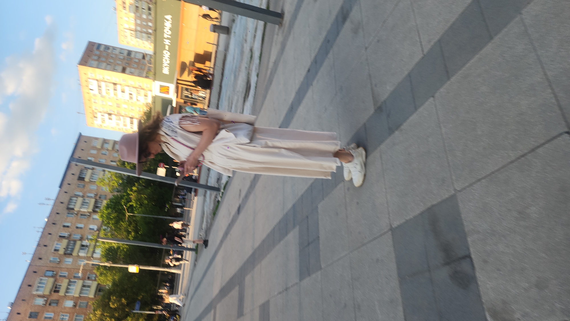A woman stands on a city sidewalk, engrossed with her mobile phone. She is dressed in a pale outfit with white shoes and a blush-colored hat. The background features typical residential buildings and a "Вкусно — и точка" restaurant, which translates to "Tasty, and that's it." The combination indicates that the scene is likely set in a modern urban area of Moscow, Russia.