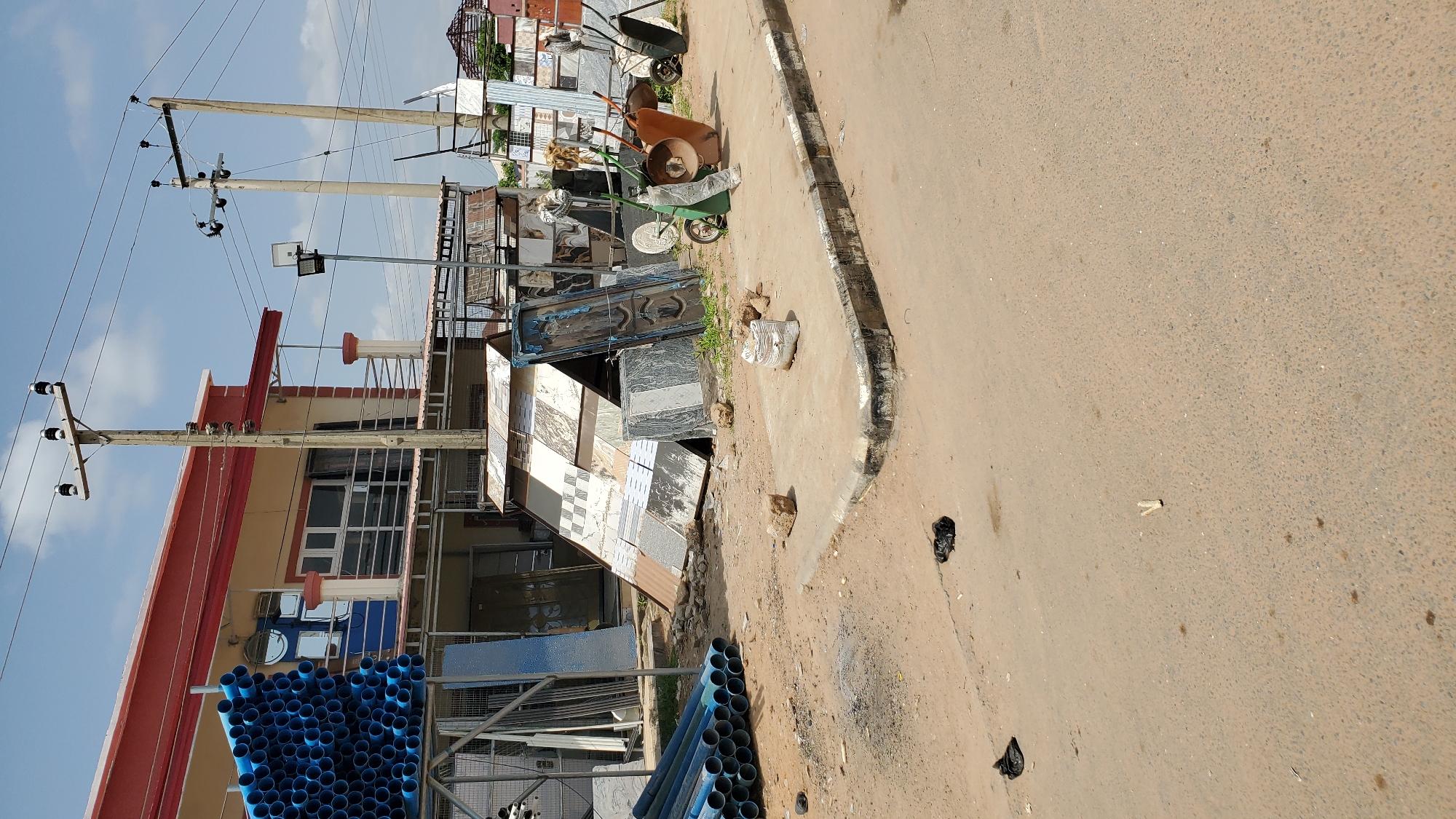 This image depicts an outdoor scene in Jalingo, Nigeria, likely a street adjacent to a building materials supplier. The time of day appears to be daytime, with bright sunlight casting shadows. The sky is partly cloudy.
The foreground is dominated by a stack of blue PVC pipes, neatly arranged. To the left, a building with a red and beige facade is visible, featuring multiple windows and some architectural details like a red railing on a balcony. Overhead, utility poles with electrical wires traverse the sky, a common sight in urban and semi-urban areas.
In the middle ground, to the right of the building and pipes, a collection of construction materials is displayed. This includes stacks of tiles with various patterns and colors, some leaning against metal racks. Several wheelbarrows, one orange and one green, are present, suggesting ongoing or recent construction activity. A dark metal structure, possibly a gate or door, is also visible among the materials.
The ground is a mix of dirt and paved surfaces, with a curb running along the edge of the paved area. Scattered debris, including what appears to be a black plastic bag and some small rocks, are visible on the ground, indicative of a less pristine environment. The overall impression is one of a functional, possibly busy, commercial area where construction supplies are sold and handled. No people are visible in the frame. There is no discernible text in the image.