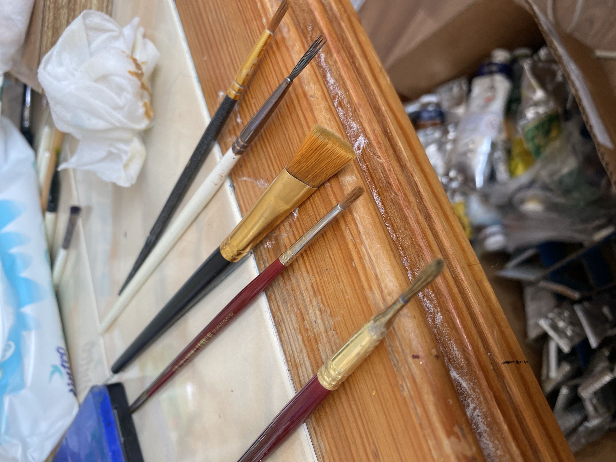 The image shows an assortment of art supplies on a wooden surface. The subjects are primarily art tools, including several paintbrushes of varying sizes and bristle types. One brush has a wide, flat head with golden bristles and a black handle. Another is a thinner brush with dark bristles and a white handle. There are also other brushes with maroon and dark handles, some with visible ferules. A crumpled white paper towel with brown stains is visible to the left, suggesting recent use or cleaning. In the background, a cardboard box is filled with what appear to be tubes of paint and other art materials. The lighting suggests it is daytime, and the setting appears to be an indoor workspace or studio. There is some white powder or dust scattered on the wooden surface. No people are visible in the image. Text is visible on the maroon brush handle: "653535362604".