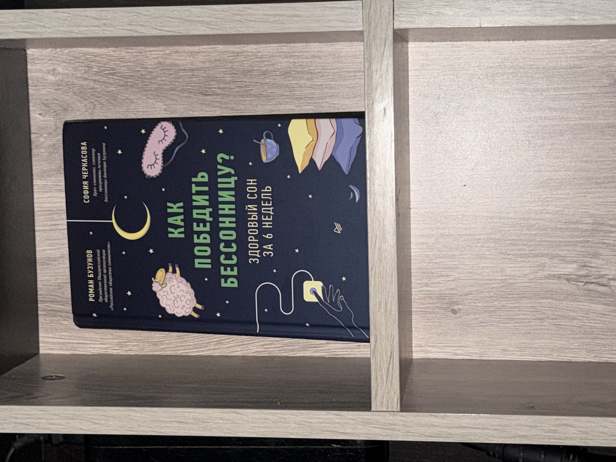 A book titled "КАК ПОБЕДИТЬ БЕССОННИЦУ?" (How to Beat Insomnia?) is displayed on a wooden shelf in a room. The book has a dark blue cover with illustrations of a sheep, a crescent moon, a sleep mask, a hand pressing a button, and a stack of pillows. The text on the cover is in Russian. The shelf appears to be part of a home or a small shop. The image was taken in Baan Mae Nam, Thailand.