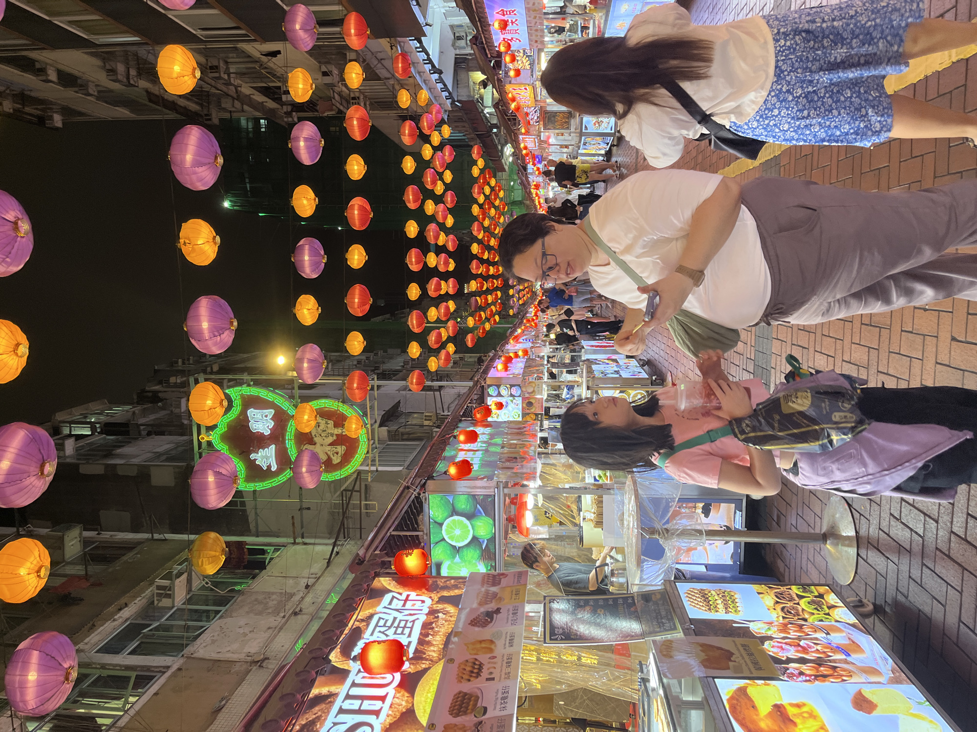 Here's a description of the image:
The photo captures a bustling night scene in Hong Kong, China, likely a street food market area adorned with numerous colorful hanging lanterns in shades of purple, orange, and red. These lanterns are strung overhead, creating a festive and lively ambiance.
On the right side of the frame, three women are seen strolling through the market. The first woman wears a pink shirt. The second wears a white shirt and gray pants, and is looking at her phone. The third woman wears a white shirt and blue floral skirt.
To the left, several food stalls display illuminated signs showcasing various culinary offerings, including waffles and other snacks. A vendor stands behind one of the stalls. The buildings lining the street are visible in the background.