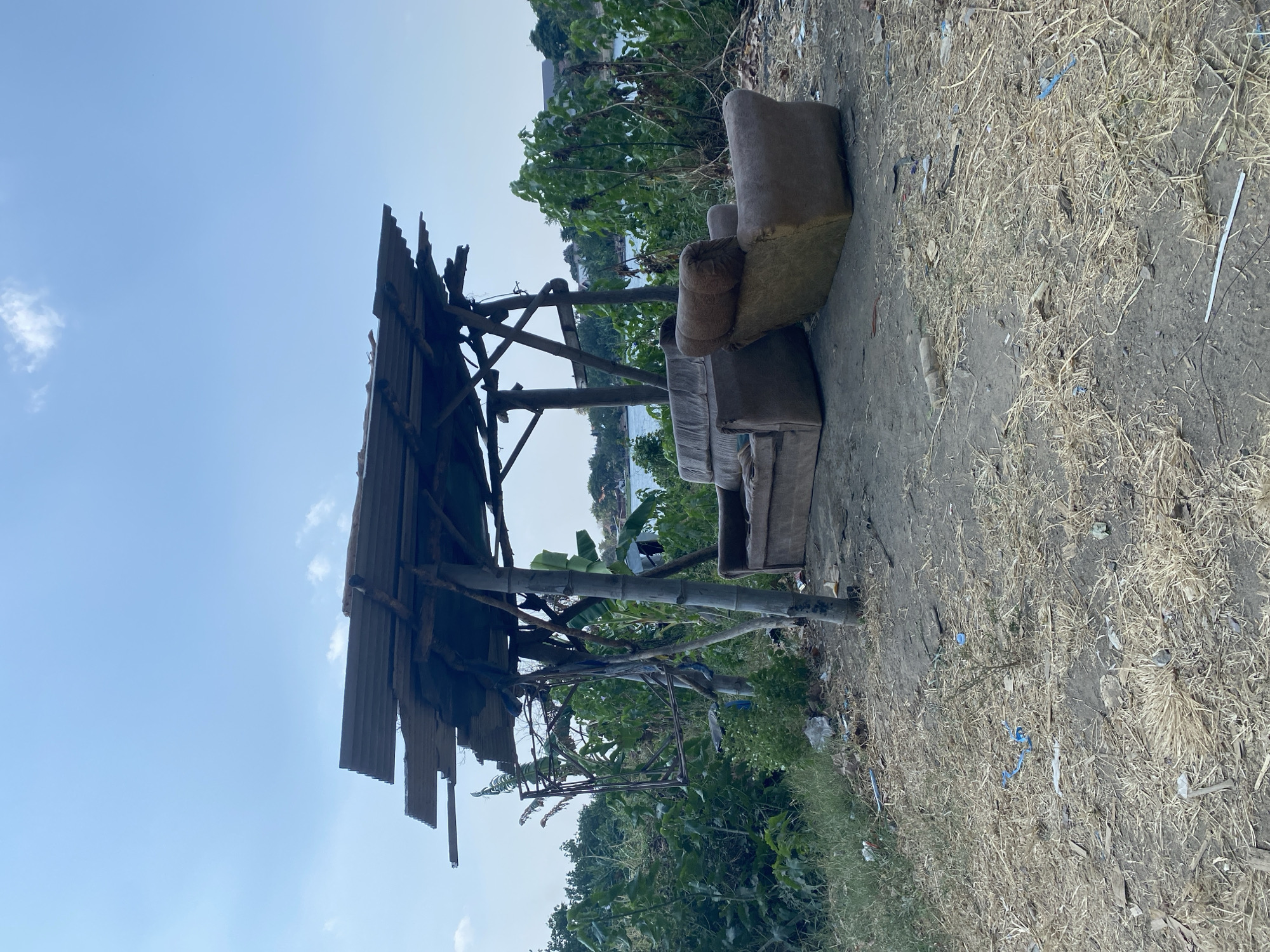 The image depicts an outdoor scene in Kediri City, Indonesia, featuring a rudimentary shelter constructed from wooden poles and corrugated metal sheets. Two worn, upholstered armchairs are placed beneath the shelter. The ground is a mixture of dry grass, dirt, and scattered debris. Beyond the shelter, lush green vegetation is visible, leading to a body of water. The sky above is a pale blue with a few wispy clouds. The overall impression is one of a neglected or improvised outdoor seating area in a natural or semi-urban environment.
