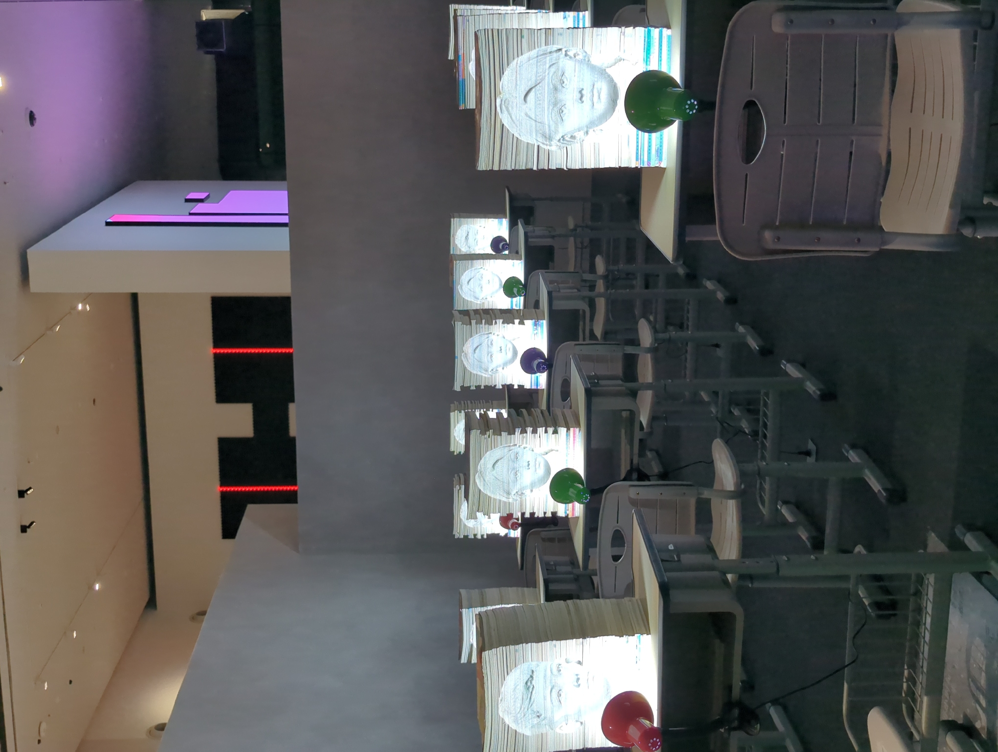 This image depicts an indoor art installation that appears to be set up in a classroom or gallery setting. The scene is dominated by rows of empty student desks, each with a stack of books on top. These books are illuminated from within, projecting the grayscale image of a child's face onto the front surface of the stack. Several colorful lamps, red and green, are placed on some of the desks, adding pops of color to the otherwise muted environment. The background shows a gray wall and a ceiling with track lighting. The overall impression is one of a dimly lit, somewhat eerie, and thought-provoking artistic display. There are no people visible in the image.
