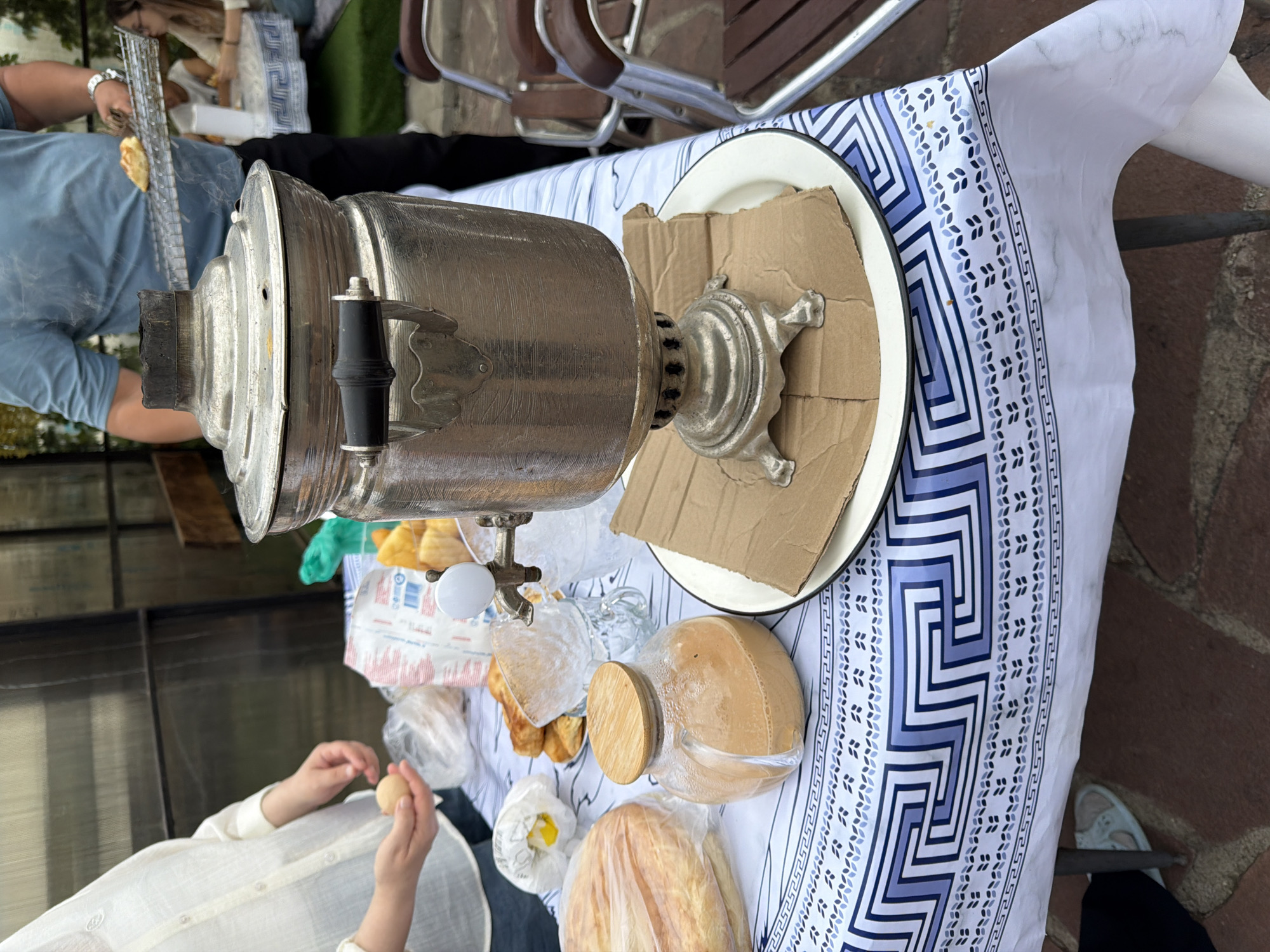 An outdoor gathering is underway, likely in Молбаза, Kazakhstan. A prominent, ornate silver samovar stands centrally on a table covered with a white and blue patterned tablecloth. Beside the samovar is a round plate with a piece of cardboard on top, and a glass jar filled with a light brown beverage. In the foreground, a large round bread, possibly lavash, is wrapped in plastic. To the left, a person in a white shirt is holding a small, light-colored object, possibly an egg. In the background, a man in a blue shirt is holding a metal rack with a pastry on it, with smoke emanating from the samovar. Other individuals are seated at a table in the distance. The setting appears to be a casual outdoor space, possibly a yard or patio, with glimpses of greenery and fencing.