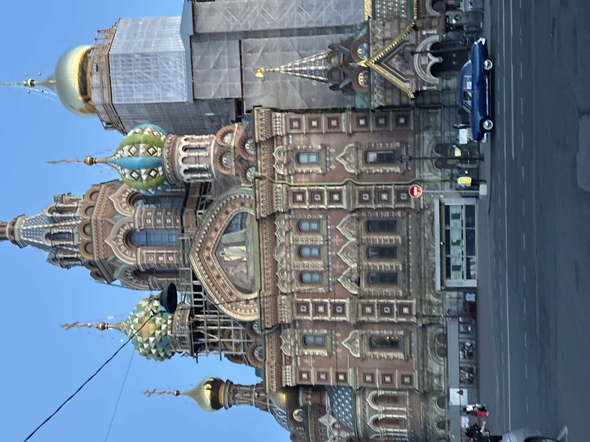 This is a medium-shot, eye-level photograph of the Church of the Savior on Spilled Blood in Saint Petersburg, Russia, taken during daylight. The sky is a clear, pale blue. The church, constructed of red brick with ornate decorative elements, features multiple domes of varying sizes and colors, including gold, green, and blue. Some of the domes are adorned with intricate patterns. Scaffolding is visible around a portion of the church, suggesting ongoing construction or restoration.
In the foreground, a blue vintage car with white-rimmed wheels is driving on an asphalt road. The car is positioned to the right of the frame, moving from left to right. A traffic light showing a red circle with a white horizontal bar is visible to the left of the car, near a lamppost. In the background, several people are walking on a sidewalk in front of the church. They are partially obscured by street furniture and the low angle of the photograph. The overall scene depicts a streetscape with a prominent architectural landmark and vehicular traffic.