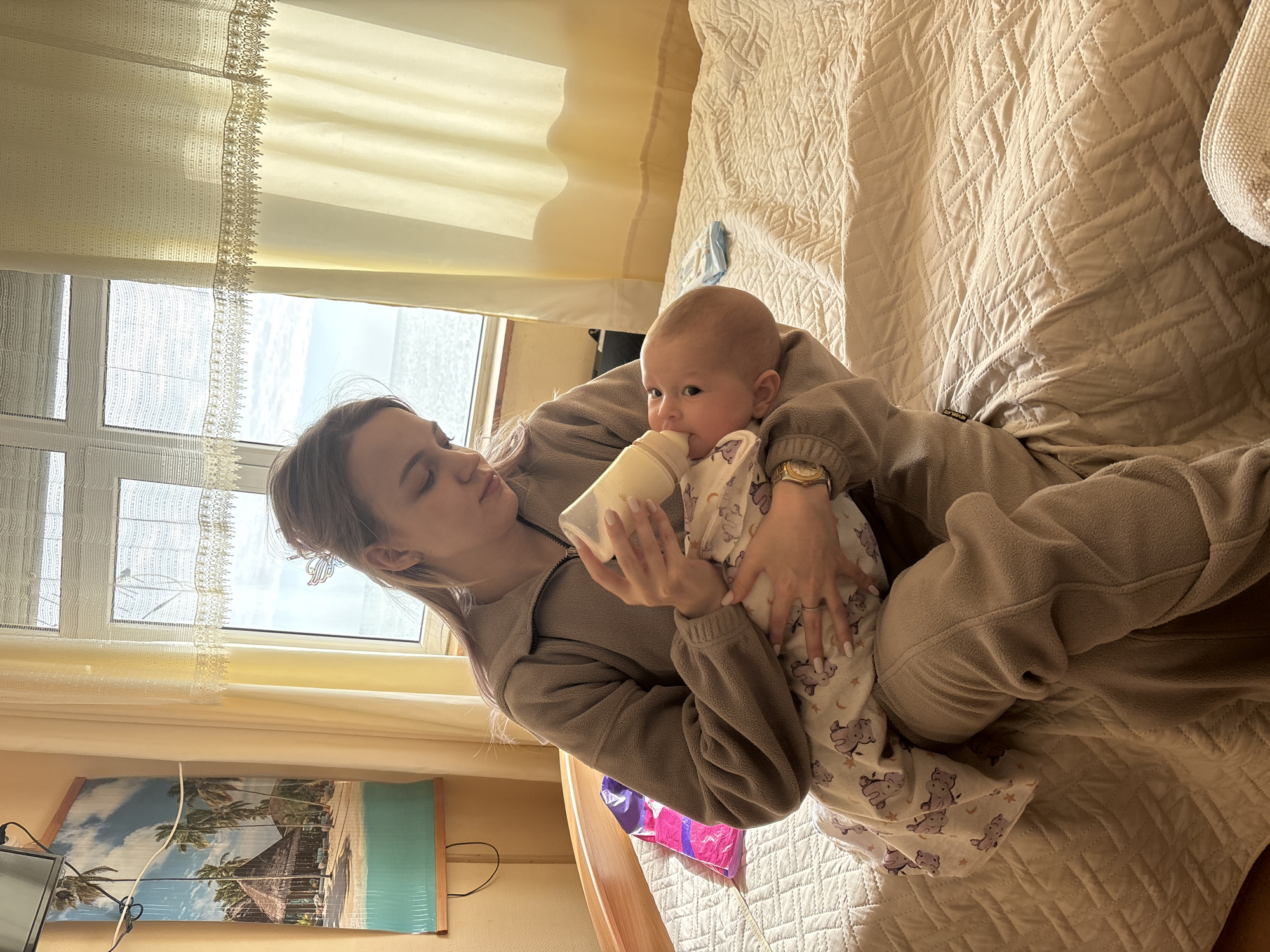 A young woman in a beige fleece zip-up jacket and matching pants is sitting on a bed in an indoor setting, holding and feeding a baby with a bottle. The baby is wrapped in a white blanket with a teddy bear print. The bed is covered with a light beige quilted blanket. Behind the woman, a window with a light-colored curtain is visible, and a landscape picture with palm trees hangs on the wall. This appears to be a personal indoor space, possibly a bedroom, and is not identifiable to the location of Sochi, Russia.