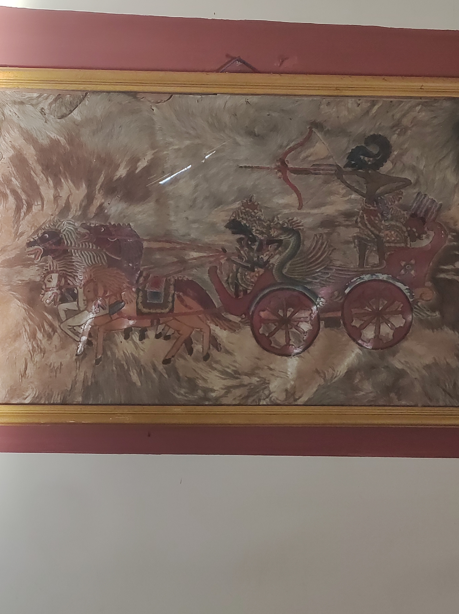 This is a painting depicting a chariot scene, likely a battle or a procession. The chariot is drawn by two horses, one white and one bay, both appearing agitated. A figure in the chariot, possibly a warrior, is aiming a bow and arrow. Another figure, partially obscured, is in the front of the chariot. The chariot itself is ornate, with a distinctive bird-like motif on the side. The background is abstract, with swirling patterns of brown and beige. The painting is framed in a gold-colored border and appears to be hanging on a wall painted a deep red. The location in Mojopuro, Indonesia, suggests it could be a historical or religious artwork.