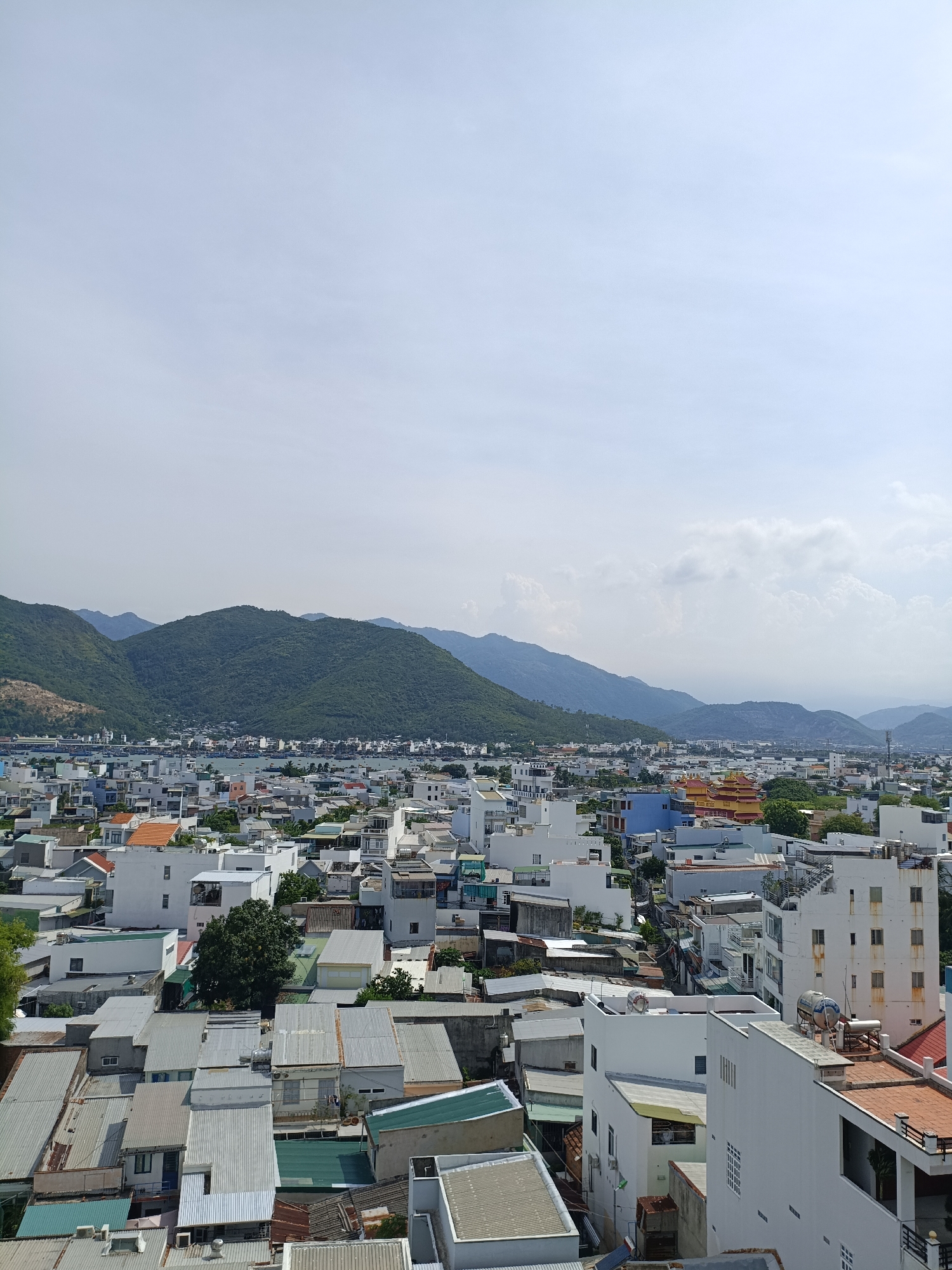 The image shows an aerial view of a densely populated urban area in Nam Nha Trang Ward, Vietnam, with a backdrop of lush green mountains. The foreground and midground are filled with a multitude of buildings of varying sizes and architectural styles, predominantly residential structures with flat roofs, many made of corrugated metal. The buildings are tightly packed together, with narrow streets and alleyways visible between them. Some buildings are painted white, while others display muted tones of orange, blue, and yellow. Rooftop structures, such as water tanks and solar panels, are common. Greenery, in the form of trees and small gardens, is interspersed throughout the urban landscape. In the distance, the imposing, verdant mountains rise, their slopes covered in dense vegetation. The sky above is a pale blue with scattered white clouds. The scene depicts a typical Southeast Asian cityscape characterized by its organic growth and the seamless integration of urban development with the natural environment.