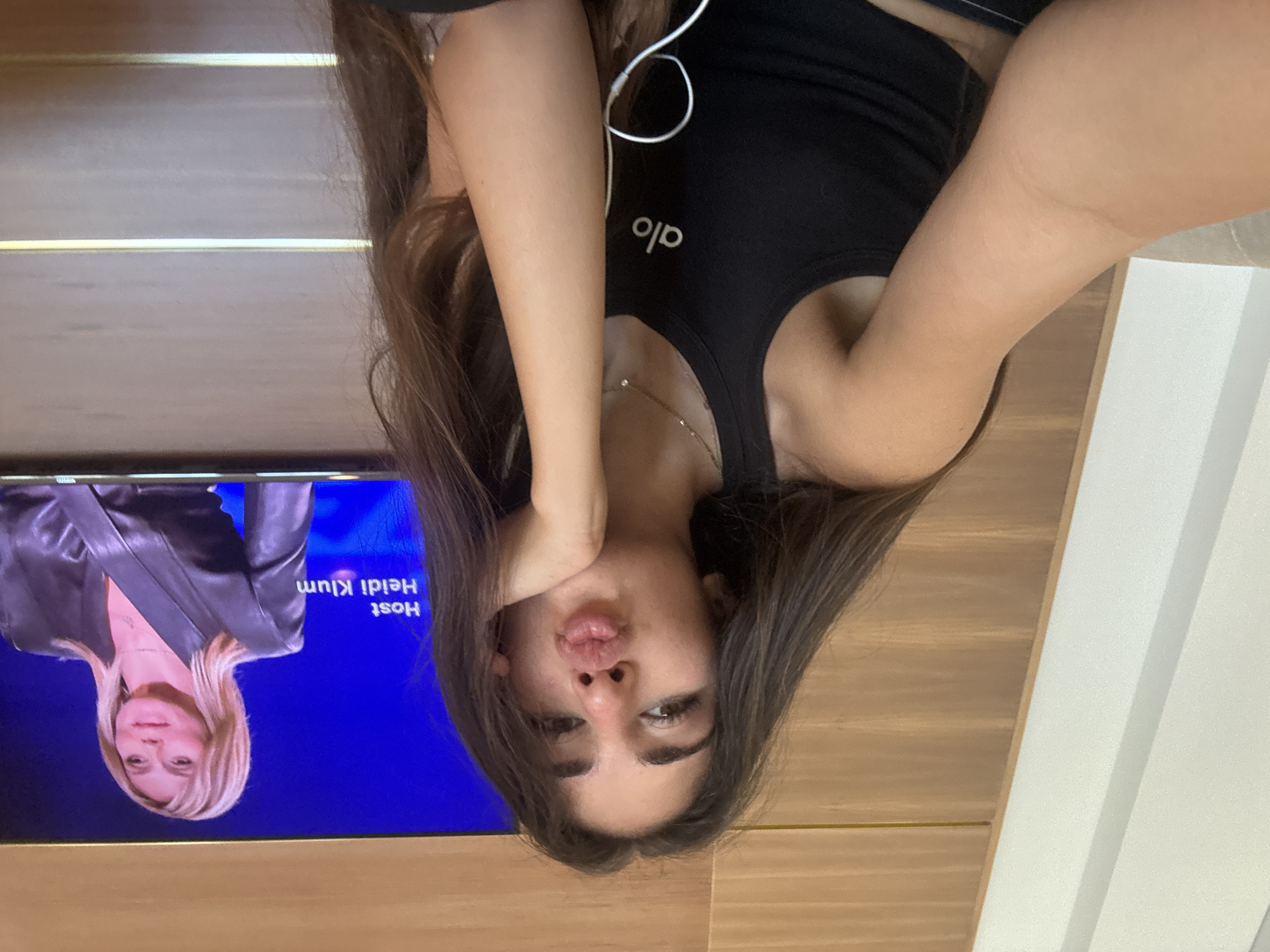 In a domestic indoor setting, a young woman with long brown hair is captured in a selfie, resting her chin on her hand and puckering her lips. She is wearing a black tank top with the "alo" logo visible. To her right, a television screen displays a segment featuring Heidi Klum, identified as the "Host," wearing a dark leather jacket. The background consists of a wall with horizontal wooden panels and a white vertical accent. The image is dated within the context of Boca Raton, United States of America.