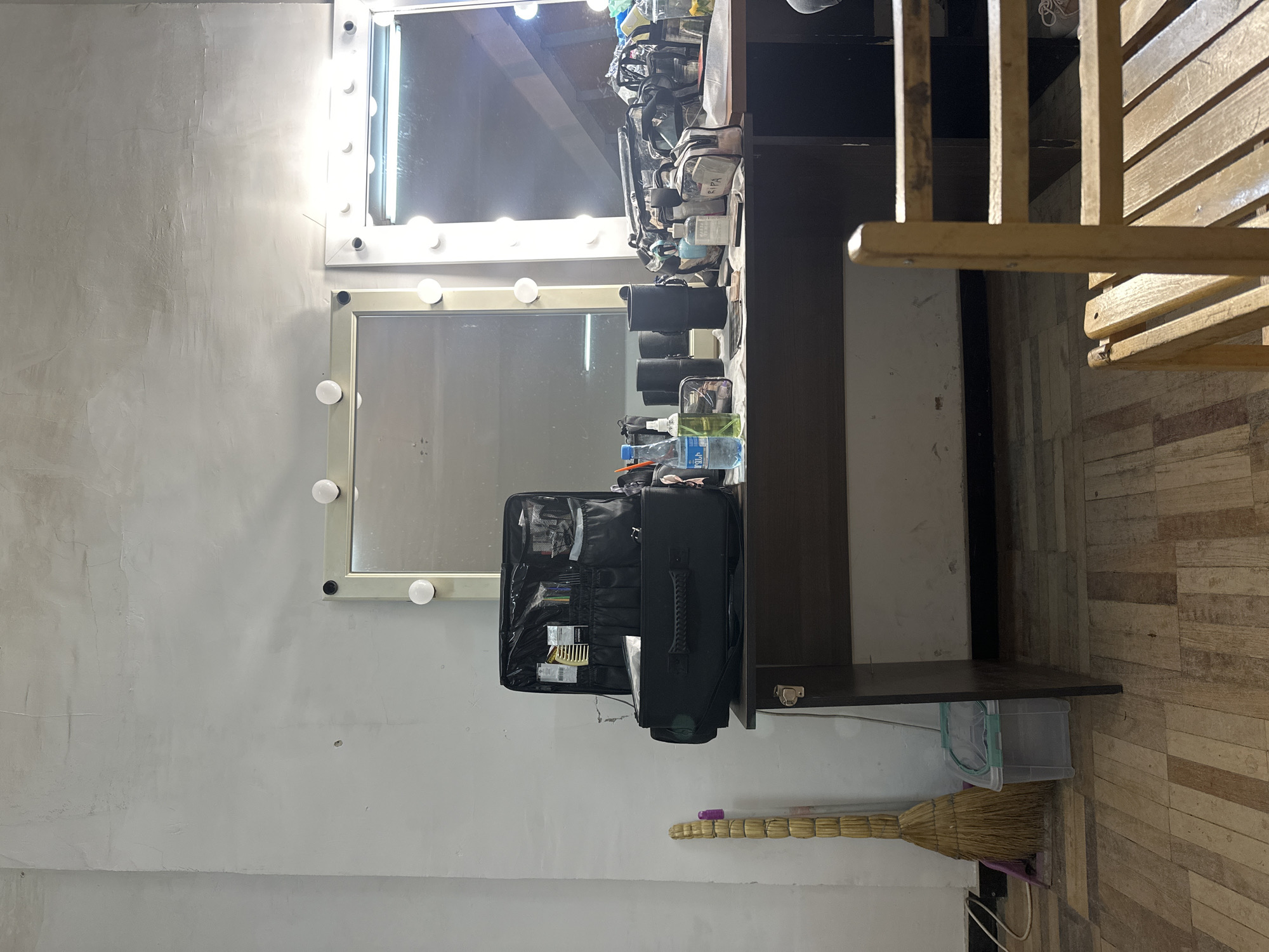 The image shows an interior space with a makeup station. The station has two mirrors with light bulbs around the frames, and a shelf below. There are makeup items and other supplies on the shelf. A black makeup bag is open on the shelf. A broom and a plastic bin are near the bottom of the image. To the right, a wooden slatted object, possibly a chair or a decorative element, is partially visible. The wall is a neutral color.
The location is Yerevan, Armenia. The image shows an interior, suggesting a dressing room or makeup studio in a building within Yerevan. There are no people visible in the image.