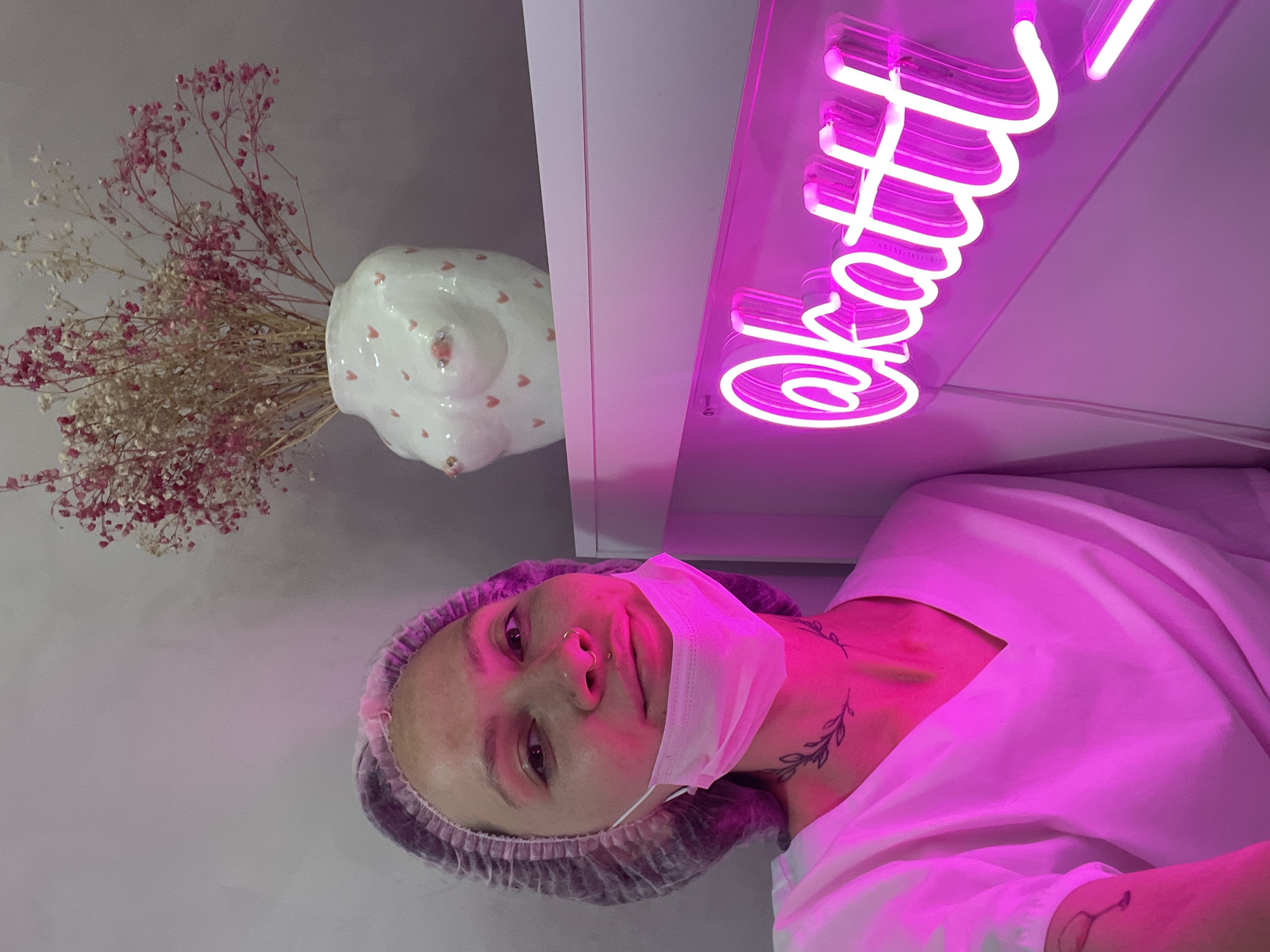 A vertical indoor shot shows a person wearing a light gray surgical cap and a disposable mask pulled down from the face. The person is also wearing a white garment. There is a tattoo on the neck and one on the wrist. In the background is a white shelf with a white vase containing dried flowers to the left. To the right is a pink neon sign reading "@katti". There is no indication of location in Izhevsk, Russia.