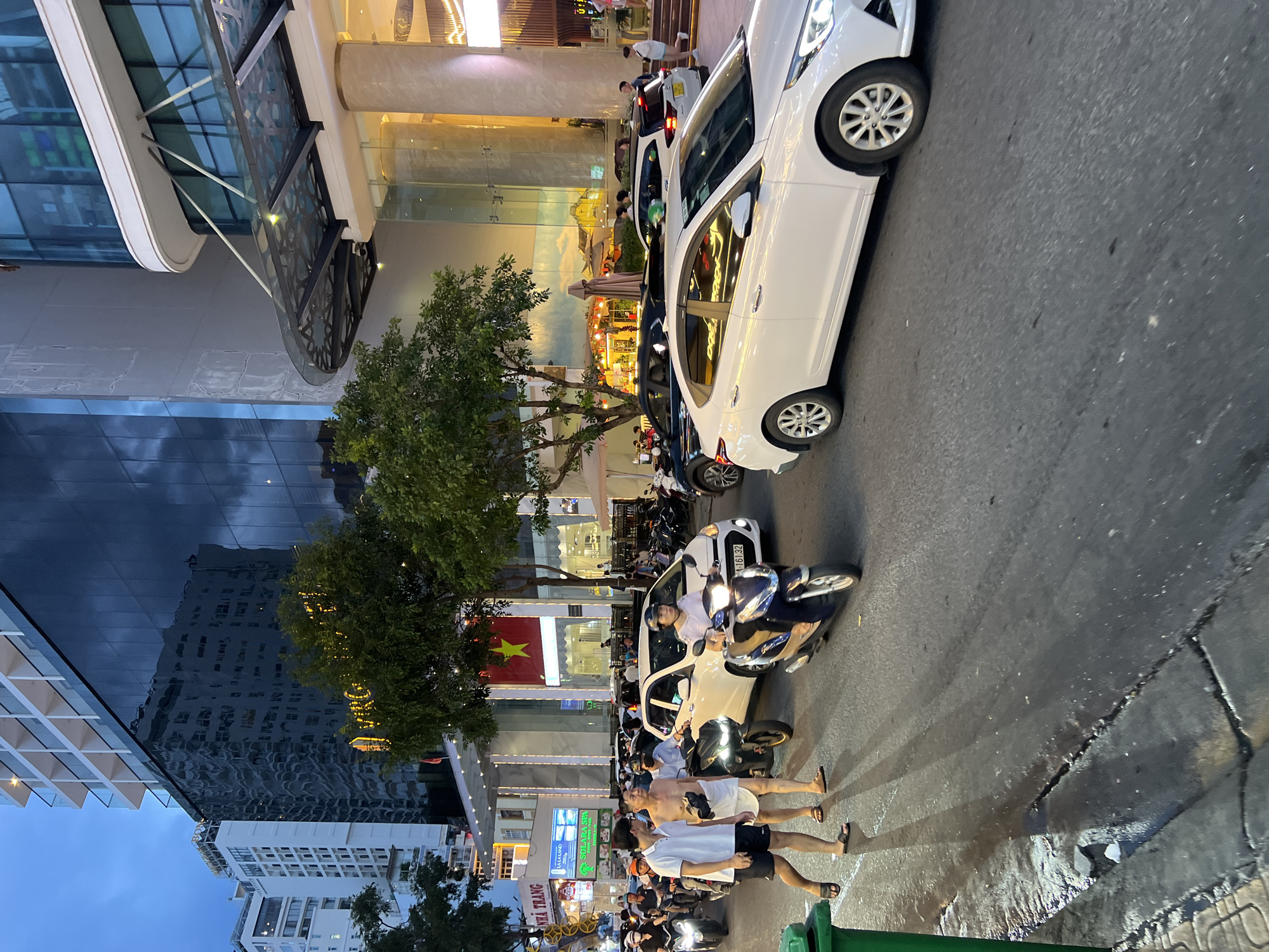 This is a medium shot of a street scene in Nha Trang, Vietnam, captured at dusk. The foreground shows a wet, asphalt road with a green garbage bin on the left. Several vehicles, including white cars and motorbikes, are navigating the street. A prominent white car with its headlights on is in the right foreground. Behind it, a blue and white motorbike with a rider is also moving. On the left side of the road, two men are walking, one wearing a white polo shirt and black shorts, and the other shirtless with a towel-like garment wrapped around his waist.
The background features a modern building with reflective dark glass and a light-colored facade. A large yellow star on a red background is visible on the building's exterior. Trees line the street, and various signs are posted, including one that reads "NHA TRANG". The overall impression is of a busy urban environment with vehicular and pedestrian traffic during the transition from day to night. The lighting suggests artificial lights are beginning to illuminate the scene.