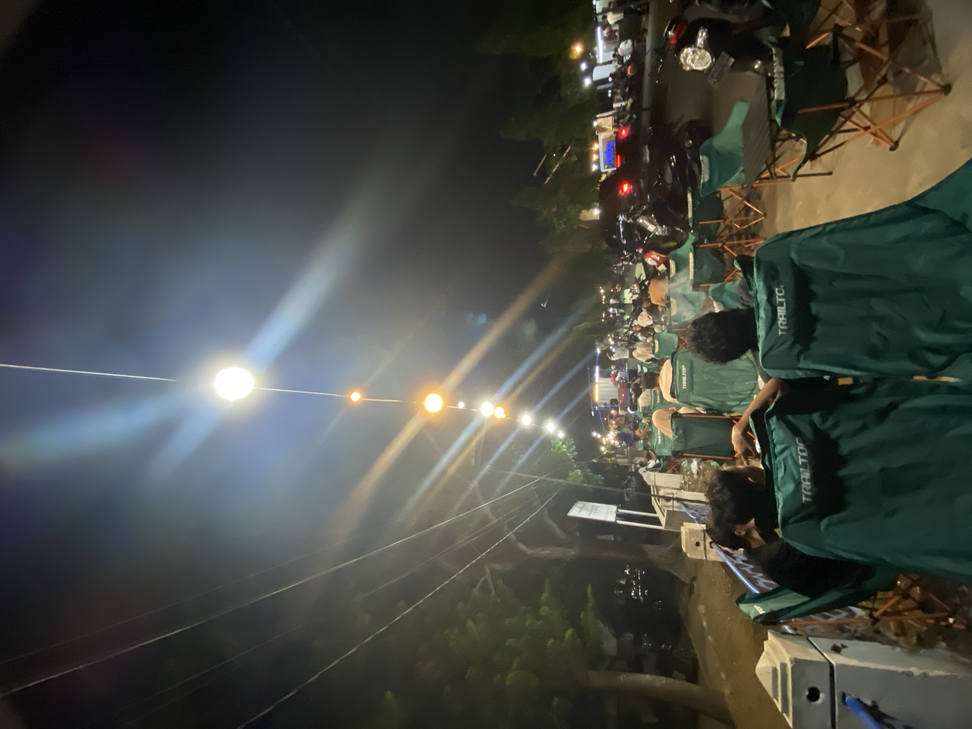 The image depicts an outdoor gathering at night in Lhokseumawe, Indonesia, likely a street-side eatery or market. Several people are seated in green camping chairs with orange frames, facing away from the camera towards a street lined with trees and illuminated by string lights and street lamps. The atmosphere is casual and social, with individuals engaged in conversation and enjoying the evening. Motorbikes and cars are parked or driving in the background, suggesting a public thoroughfare. The lighting creates lens flares and a somewhat hazy effect, common in nighttime photography with bright light sources. The foreground features two large green chairs prominently, with the word "TRAILTOP" visible on the backrests. The overall scene conveys a sense of community and a lively evening atmosphere in the city.