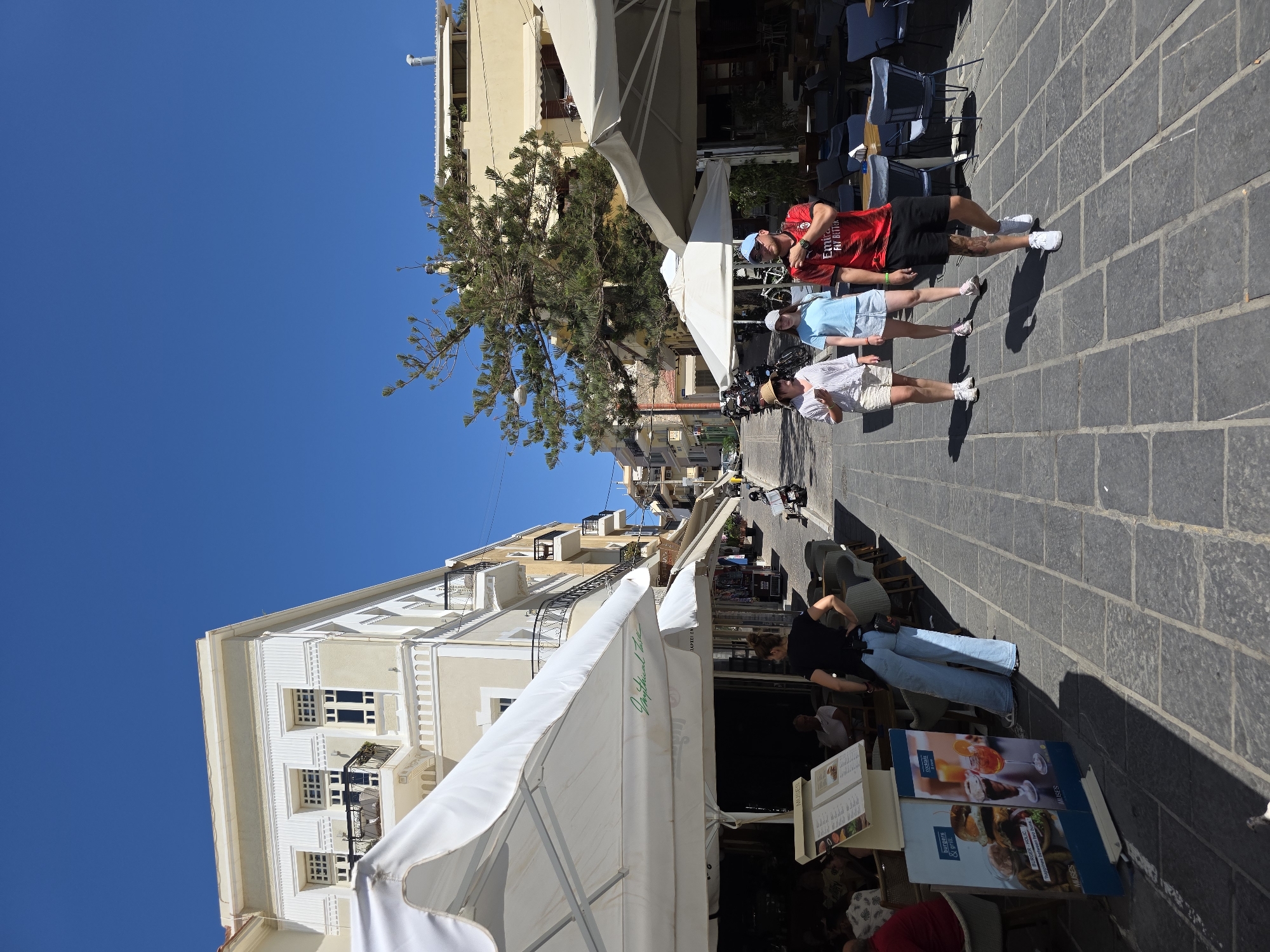 The image depicts a sunny day on a cobblestone street in Chania, Greece, likely in the Old Town, characterized by the Venetian architecture. In the foreground, a woman in a black top and light blue jeans stands with her hands on her hips, facing away from the camera, possibly a server. To her left, a menu board displays "Burgers & Grill" and "Cocktails." Large white umbrellas provide shade over outdoor seating areas of a cafe or restaurant. Further down the street, three individuals walk towards the viewer: two women in casual summer attire and a man in a red and black soccer jersey, black shorts, and a blue baseball cap. The street is lined with buildings with balconies and shops, and several motorcycles are parked on the side. The sky is a clear, bright blue.