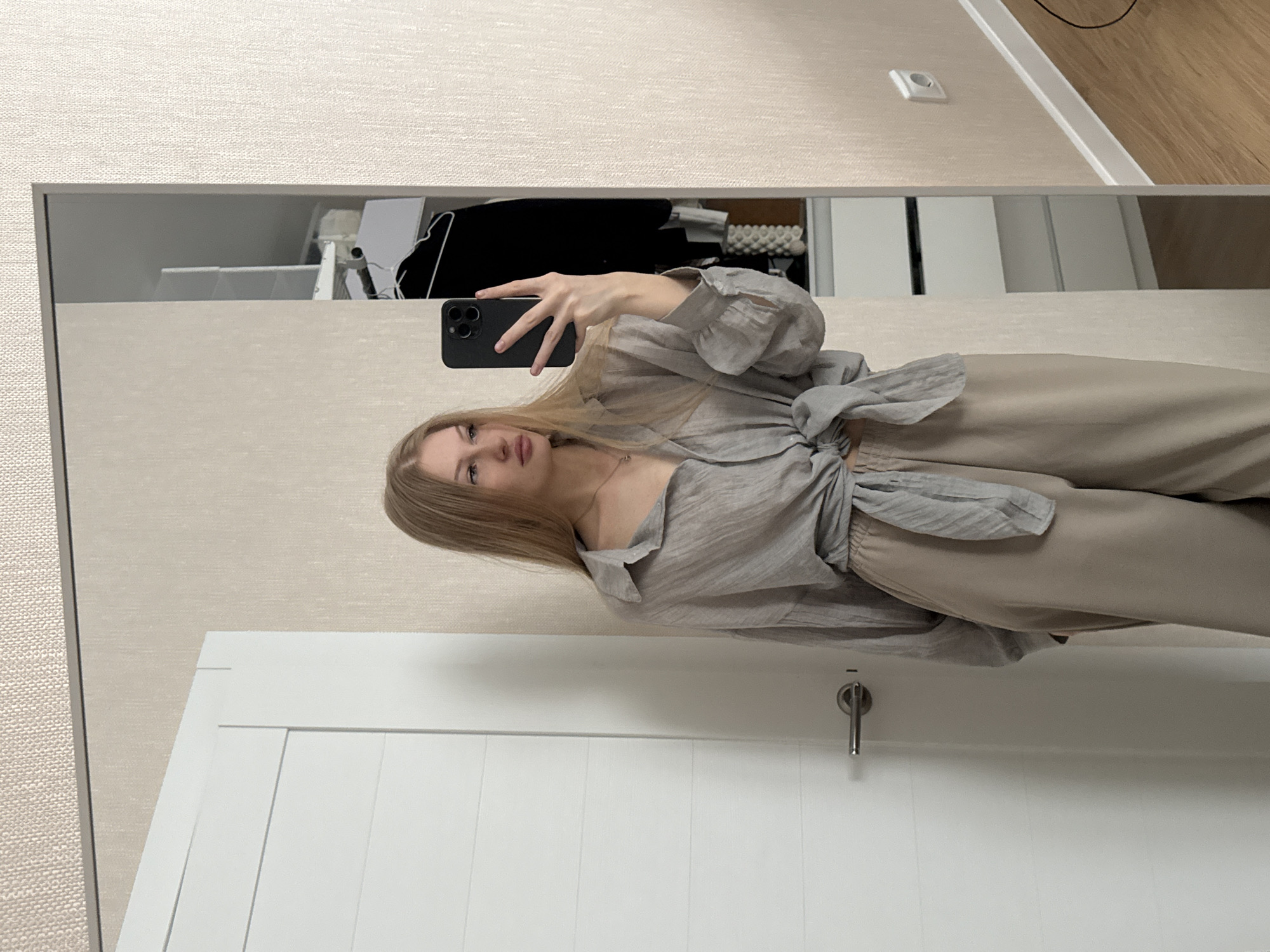 A young woman with blonde hair takes a mirror selfie in a room. She is wearing a grey, oversized, button-down shirt tied at the waist and light brown wide-leg pants. She holds a black smartphone in her right hand. Behind her, a white door is visible on the left, and to the right, a closet with clothes hanging on hangers is reflected in a mirror. The wall in the background has a textured wallpaper. The scene appears to be indoors, likely in a residential setting. The location is identified as the city of Perm, Russia.