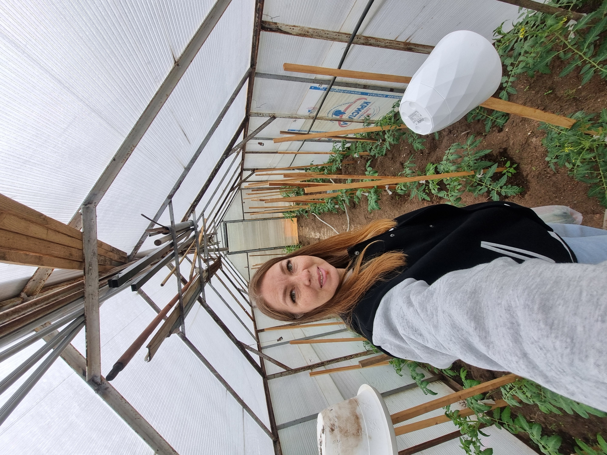 A woman stands inside a greenhouse in Chaikovsky, Russia. The greenhouse has a metal frame with a corrugated plastic roof and walls. Wooden slats run along the sides of the interior. Plants grow in the soil beneath the slats, including young tomato plants with wooden stakes to support them. Two overturned white plastic pots sit among the plants. The woman has long, reddish-brown hair and wears a black jacket with white stripes. She faces the camera and smiles.