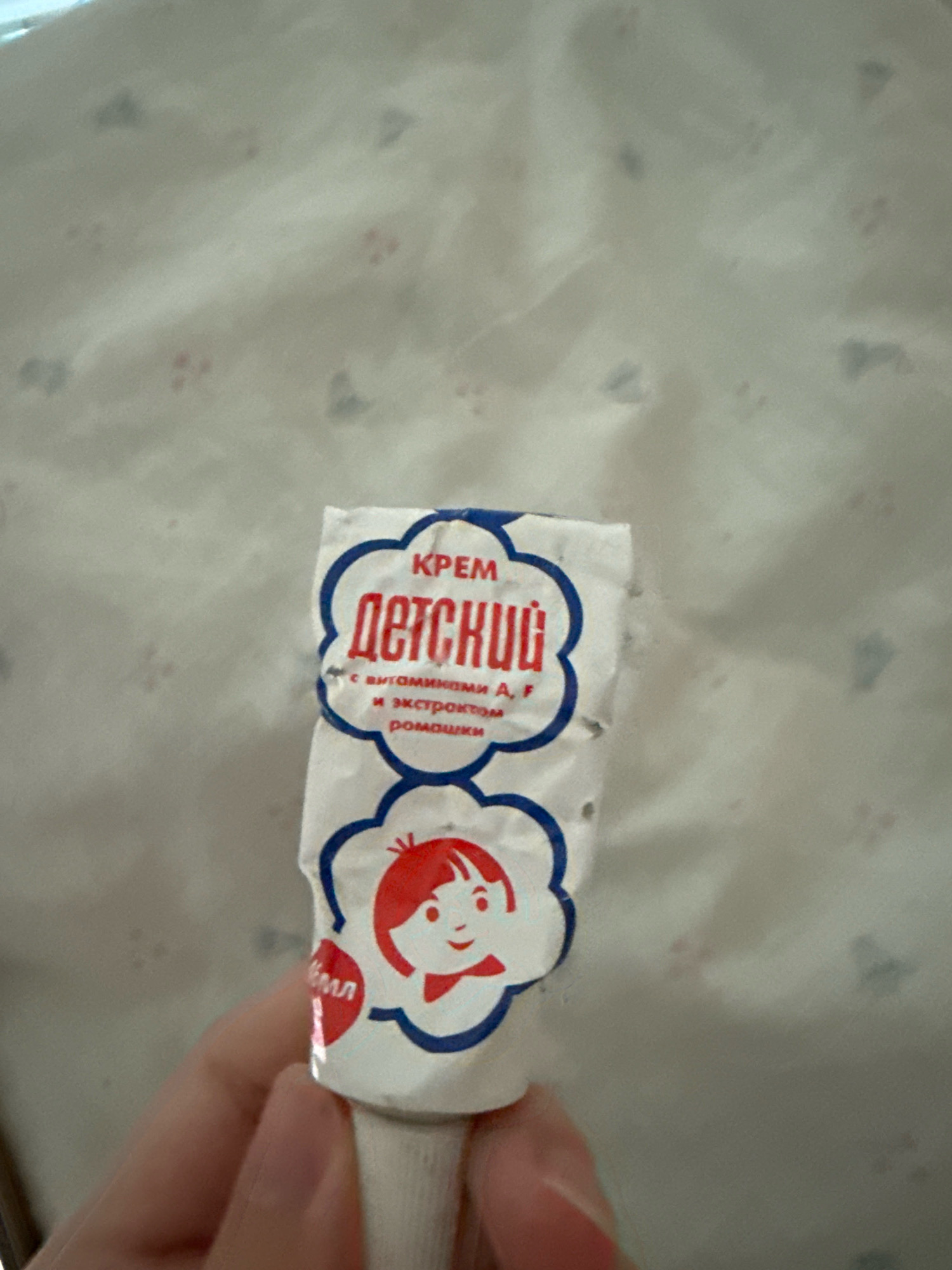 A hand holds a small, crumpled tube of "ДЕТСКИЙ" (Children's) cream. The tube is white with a blue outline and features a cartoon drawing of a smiling child with red cheeks and a red bow. The text on the tube is in Russian and indicates the cream contains vitamins A, F, and chamomile extract. The background is a light-colored fabric with a subtle pattern of small, scattered shapes. The image appears to be taken indoors in Zavolzhye, Russia.