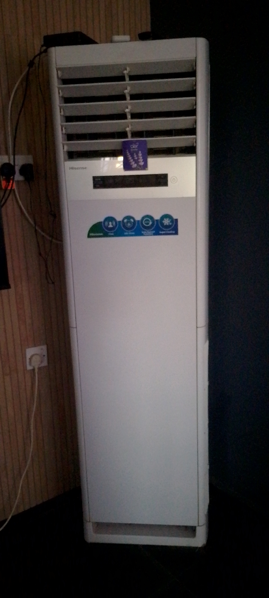 An indoor shot shows a tall, white Hisense brand floor fan with adjustable louvers on top. A small, purple sticker with a floral design is affixed to the louvers. Below the louvers is a black digital display panel and a circular button. Further down the front of the fan, a green and blue sticker displays four circular icons with text underneath, likely representing different functions. To the left of the fan, against a wood-paneled wall, a white electrical cord connects to a wall socket with a white plug. Another black cord extends from the top of the fan and runs behind it. The fan is positioned on a black floor, with a dark blue wall or surface to its right. The lighting is dim, suggesting an indoor environment.