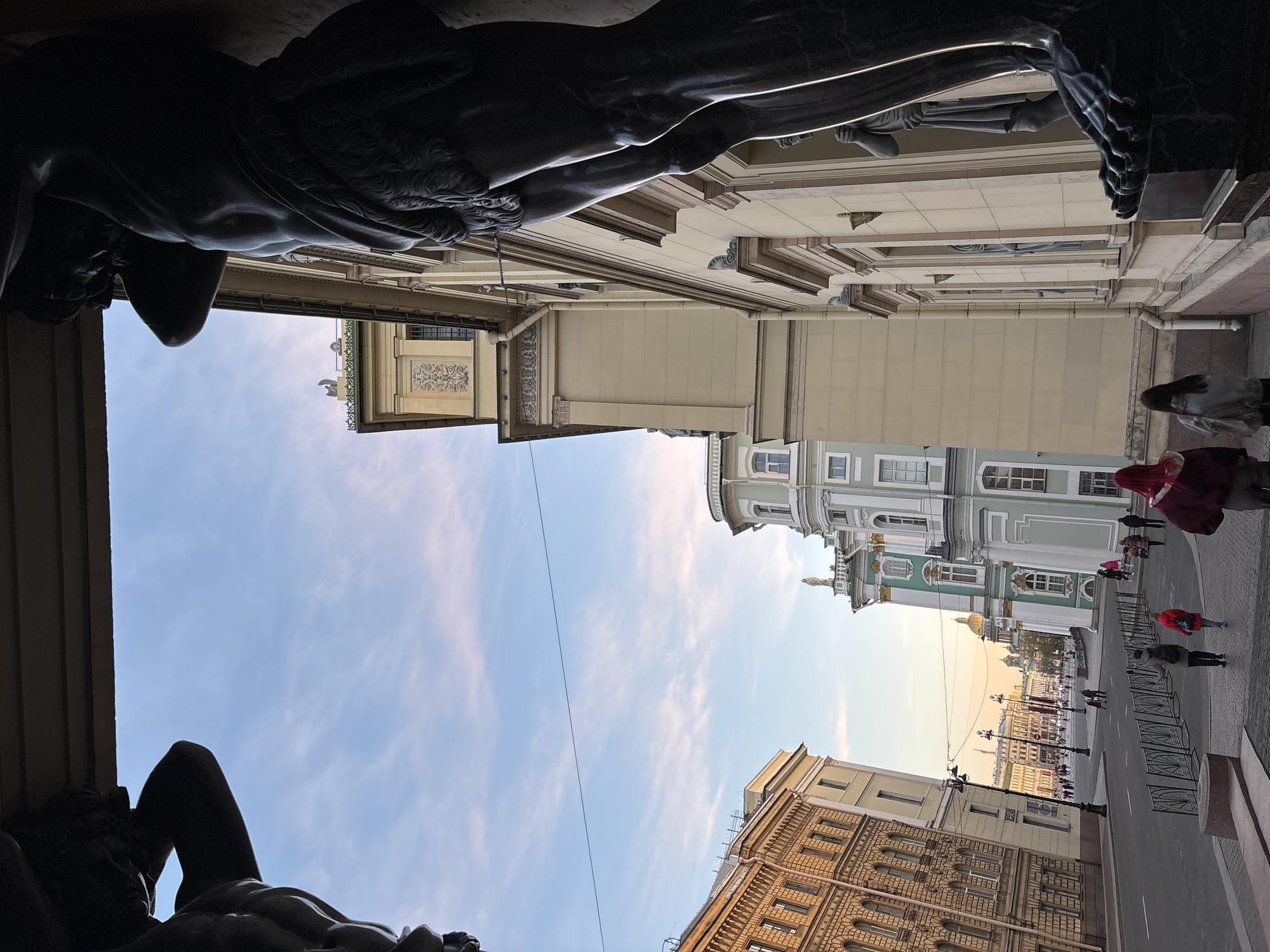 The image shows a view upward from beneath the portico of the New Hermitage Museum in Saint Petersburg, Russia. Two of the museum's iconic Atlantes sculptures, which support the portico's roof, are visible in dark silhouette at the top and bottom of the frame. Between the Atlantes, the photograph captures a view of the sky and the surrounding architecture. The State Hermitage Museum and Winter Palace are visible. The sky is light blue with wispy clouds. On the ground level, pedestrians, some in red outerwear, are seen walking along the street. There are metal barricades lining the street. A power line stretches across the sky between the buildings. The overall atmosphere is one of a bright, sunny day in a historic urban setting.