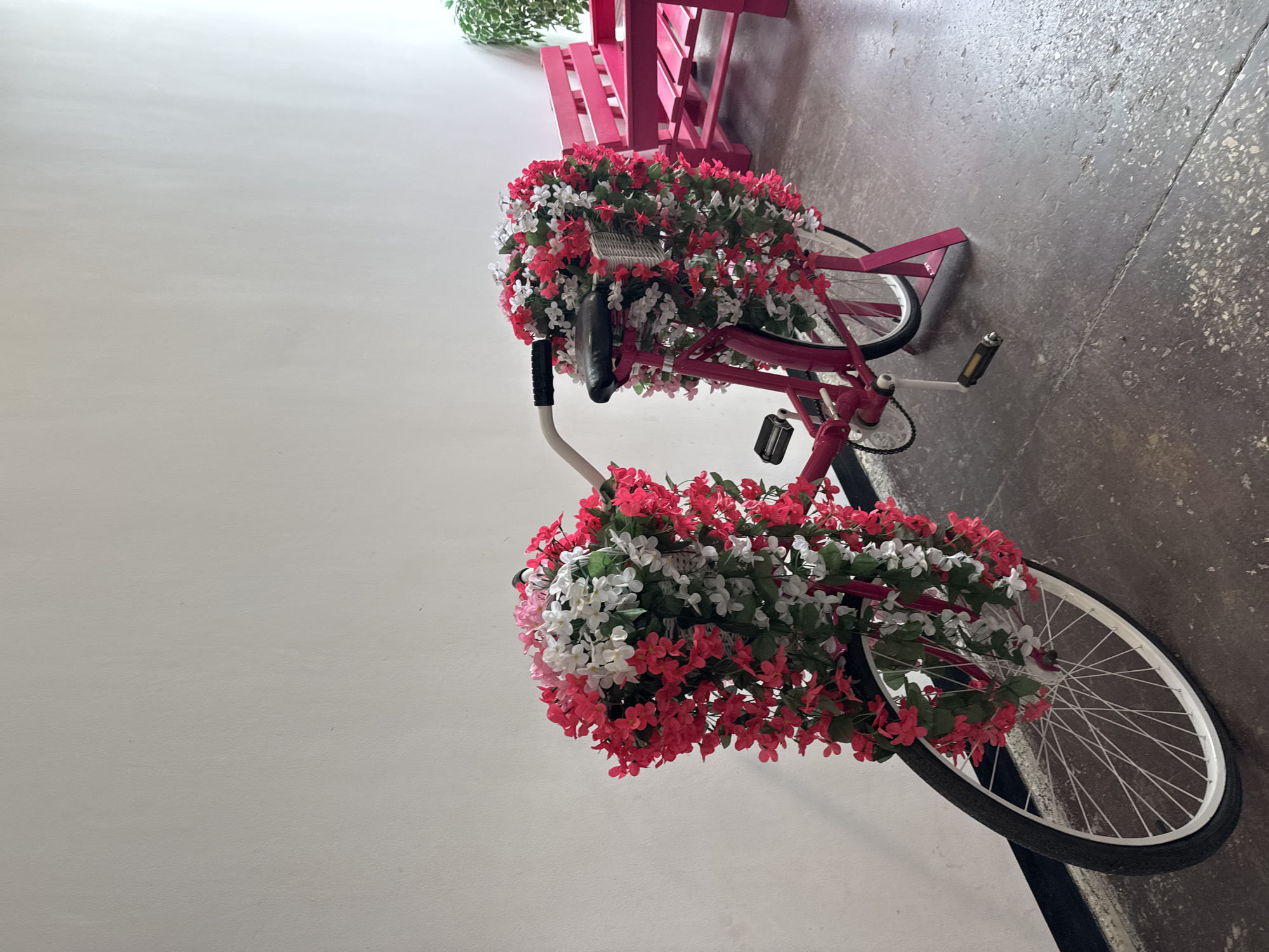 A pink bicycle decorated with red and white artificial flowers is parked on a gray concrete floor next to a white wall. The bicycle has a basket on the front, also filled with flowers. A pink bench is visible in the background, and to the right, a potted green plant. The scene is indoors, possibly in a covered walkway or an alcove. The lighting suggests it is daytime.