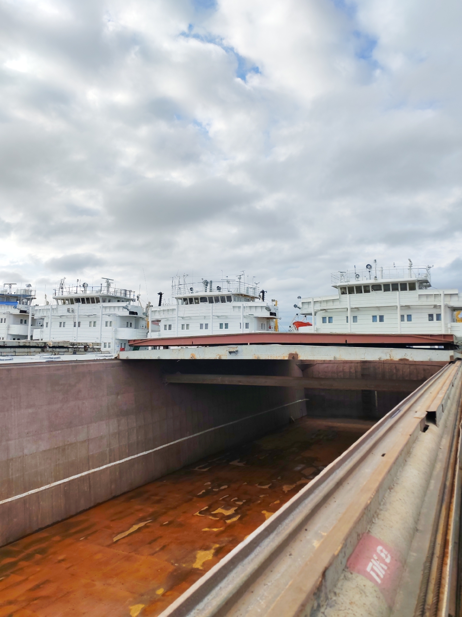 The image depicts a barge docked in Rybinsk, Russia. The barge is rusty and appears to be empty. Three white ships with multiple windows and red trim are docked next to the barge. The sky is overcast.
