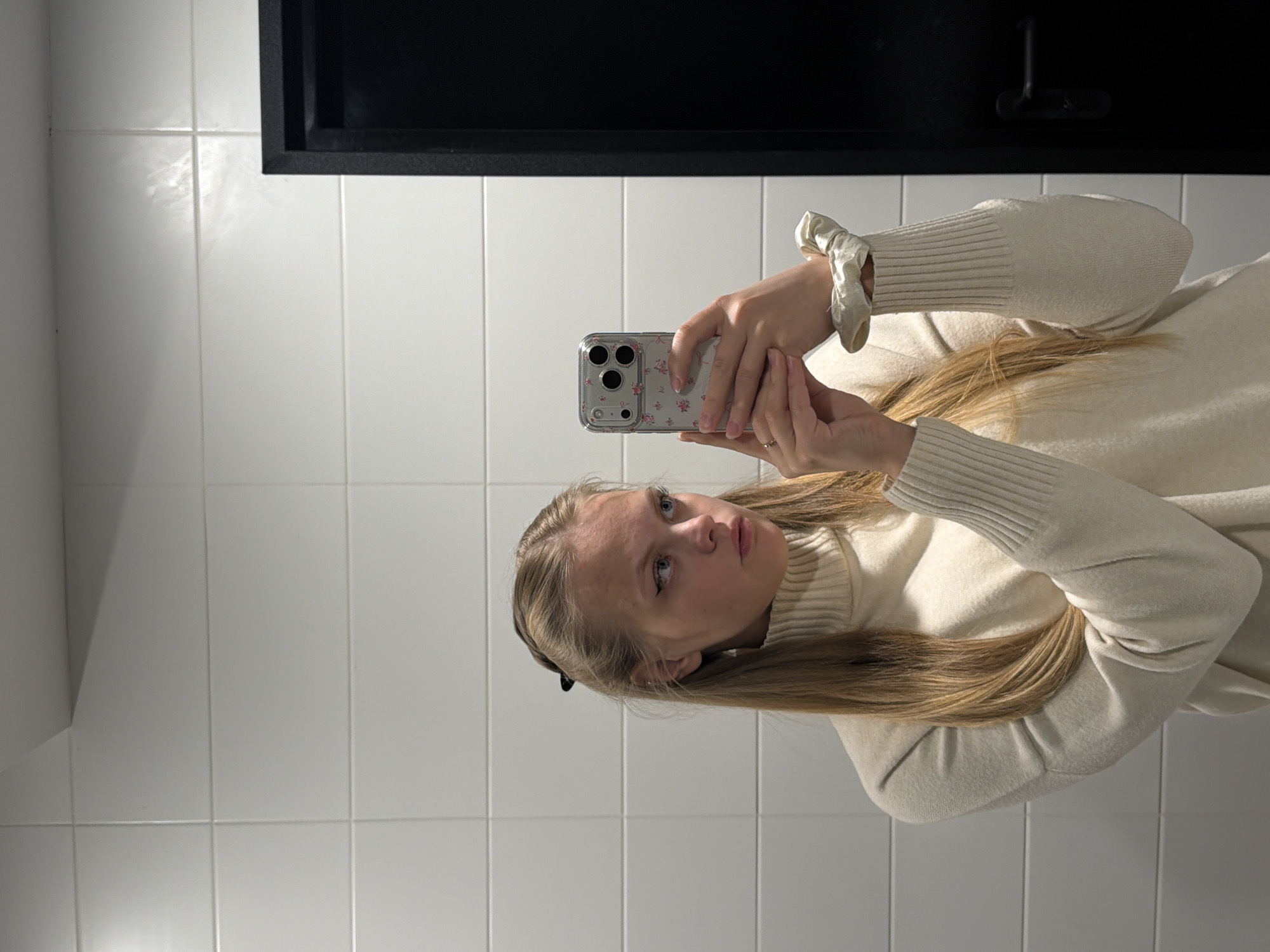 A woman is taking a selfie in a bathroom. The bathroom has white tiled walls and a dark rectangular feature above. The woman is wearing a cream-colored turtleneck sweater with ribbed sleeves and a scrunchie on her left wrist. She is holding a smartphone with a clear case decorated with small pink flowers. The location is not identifiable as Izhevsk, Russia.