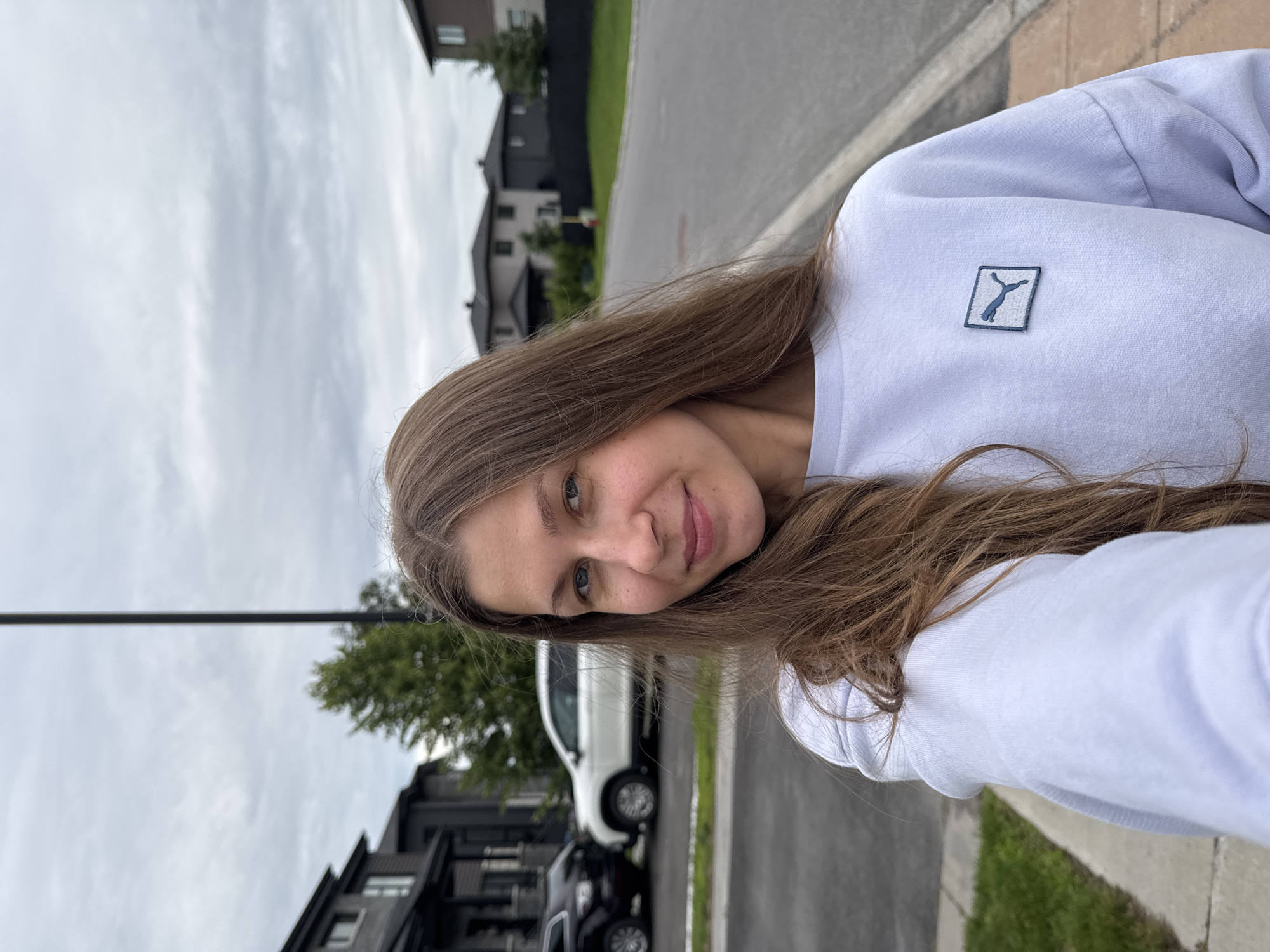 A young woman with long, brown hair is taking a selfie outdoors on a cloudy day. She is wearing a light purple sweatshirt with a small, blue Puma logo on the left chest. The woman is smiling slightly and looking at the camera. In the background, a suburban street is visible, lined with modern houses. Two cars, a black SUV and a white SUV, are parked on the side of the road. The environment appears to be a residential area in Gatineau, Canada, based on the architectural style of the houses and the overall landscape. The ground is paved with asphalt, and there are sidewalks and grass areas. The sky is overcast, suggesting a temperate climate.