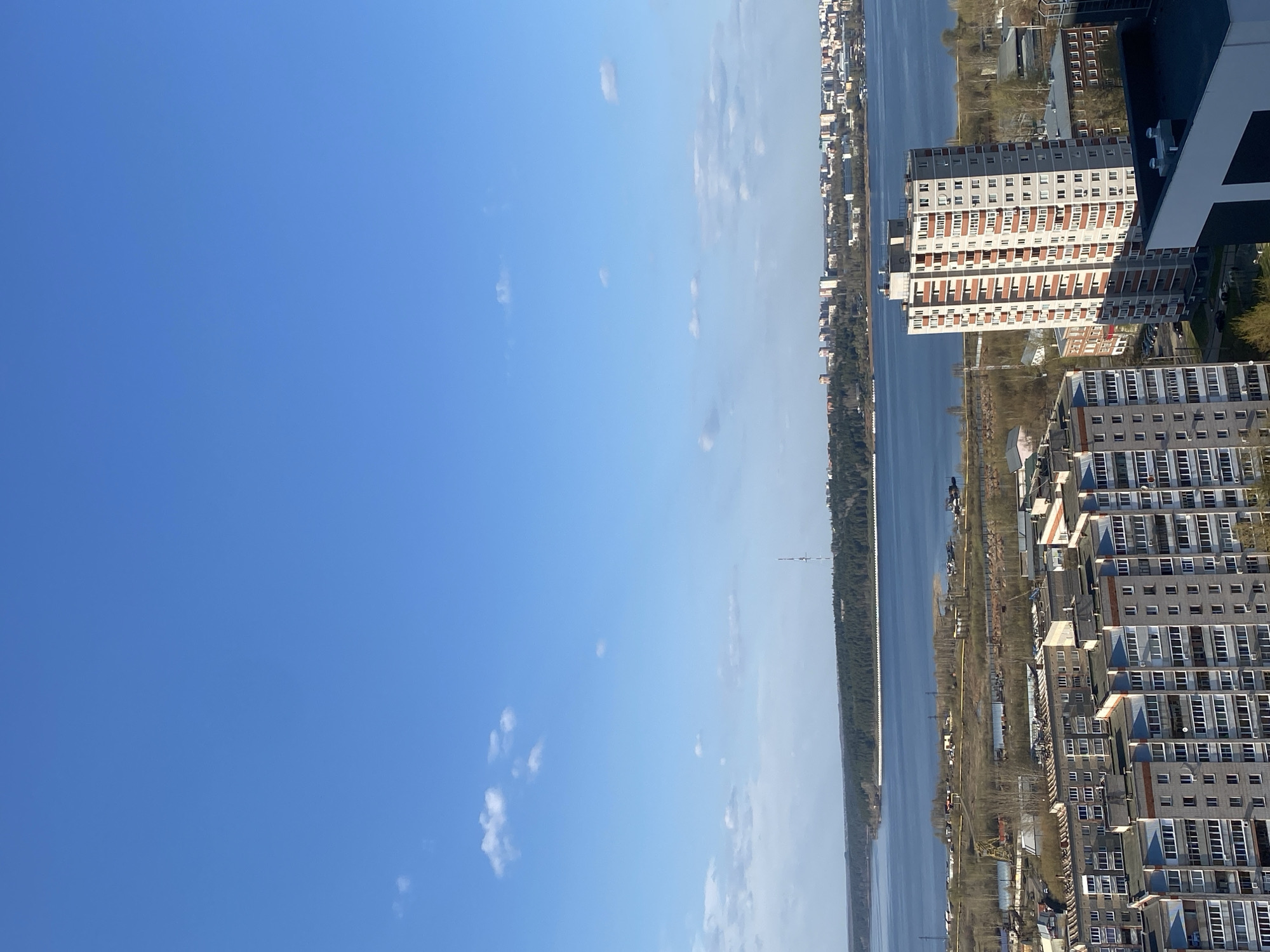 An aerial view from a high vantage point over the city of Izhevsk, Russia, features a clear blue sky with scattered cirrus clouds filling the left half of the frame. The right half is dominated by the city infrastructure, divided by a wide river or body of water. Along the river, dense tree lines appear.
Closer to the viewer, tall residential buildings rise from a developed urban area. One building, partially cropped at the upper right, has white and dark gray exterior elements with numerous windows. Below it stands a taller building with alternating rows of red-brown and white brick. Further down, several gray residential buildings with numerous windows form a cluster.
On the opposite bank, a dense row of buildings extends along the river, gradually diminishing in size as they stretch into the distance. The scene is bathed in bright daylight, suggesting a clear and sunny day.