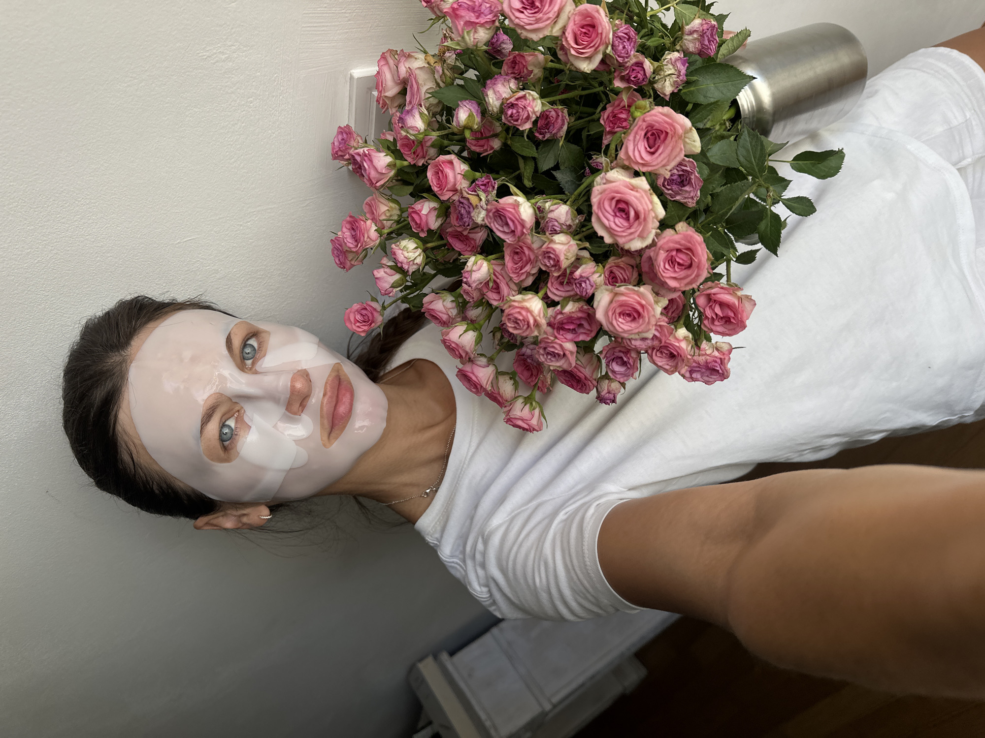The image shows a young woman with a facial sheet mask on, holding a large bouquet of pink roses in a silver vase. She is wearing a white t-shirt. The background is a plain, light-colored wall. The lighting suggests an indoor setting, possibly a home. There is no indication of the city of Boreti, Montenegro.