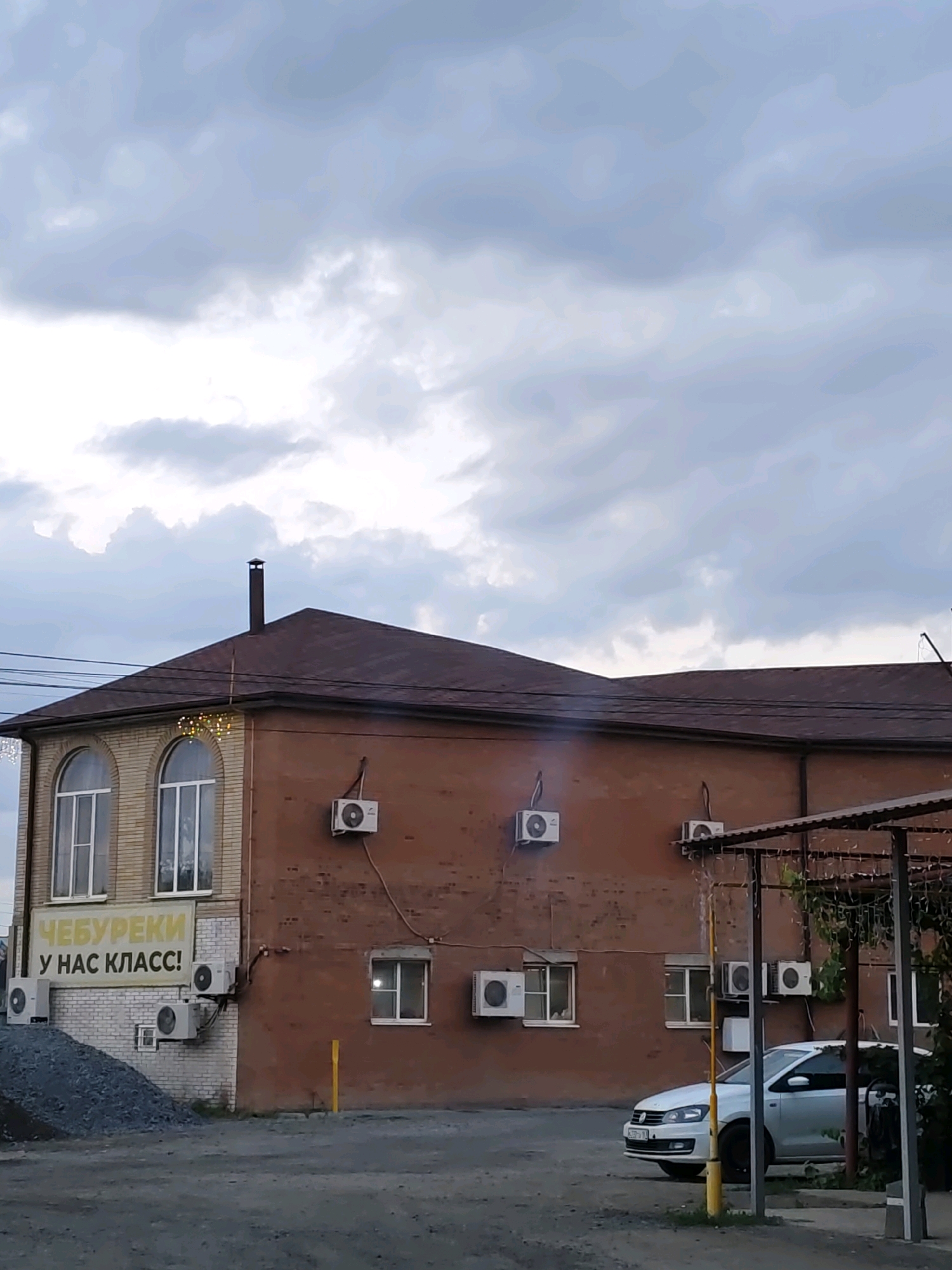 The image depicts a two-story building with a brown brick facade, situated in Rostov, Russia. The building has a dark brown, sloped roof. Multiple air conditioning units are visible mounted on the exterior walls of both floors. The upper floor features arched windows with white frames. A sign on the lower part of the building's left side, near the entrance, displays yellow text on a white background that reads "ЧЕБУРЕКИ У НАС КЛАСС!" which translates to "Chebureki are class!"
In front of the building, a white car is parked. The foreground consists of a gravel or asphalted area. The sky above is overcast with heavy clouds, suggesting a cloudy or possibly stormy day. The overall impression is of a commercial establishment, likely a cafe or eatery, in an urban setting.
