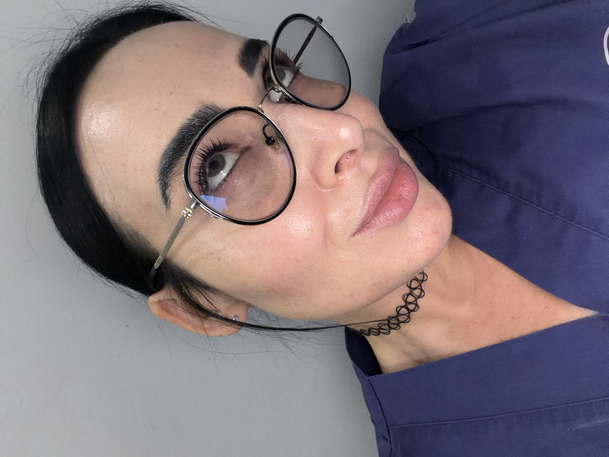 A close-up, diagonally oriented portrait features a person, likely female, captured from the chest up. The individual has dark hair pulled back from her face, prominent dark eyebrows, and long eyelashes. She wears round glasses with thin metal frames, the lenses of which reflect light with subtle blue and purple tones. Her lips are slightly parted in a soft pout, and her complexion is smooth with some visible skin texture on her forehead and cheeks.
She is dressed in a dark blue garment, possibly a uniform or scrub top, and accessorizes with a distinctive black, woven "tattoo" choker necklace around her neck. The subject is looking upwards and slightly to the right, conveying a serene or contemplative expression.
The background is a plain, light grey wall, suggesting an indoor setting with consistent, bright lighting. No specific activity beyond posing for the photograph is discernible. There is no visible text in the image. The location context for this media is Saint Petersburg, Russia.