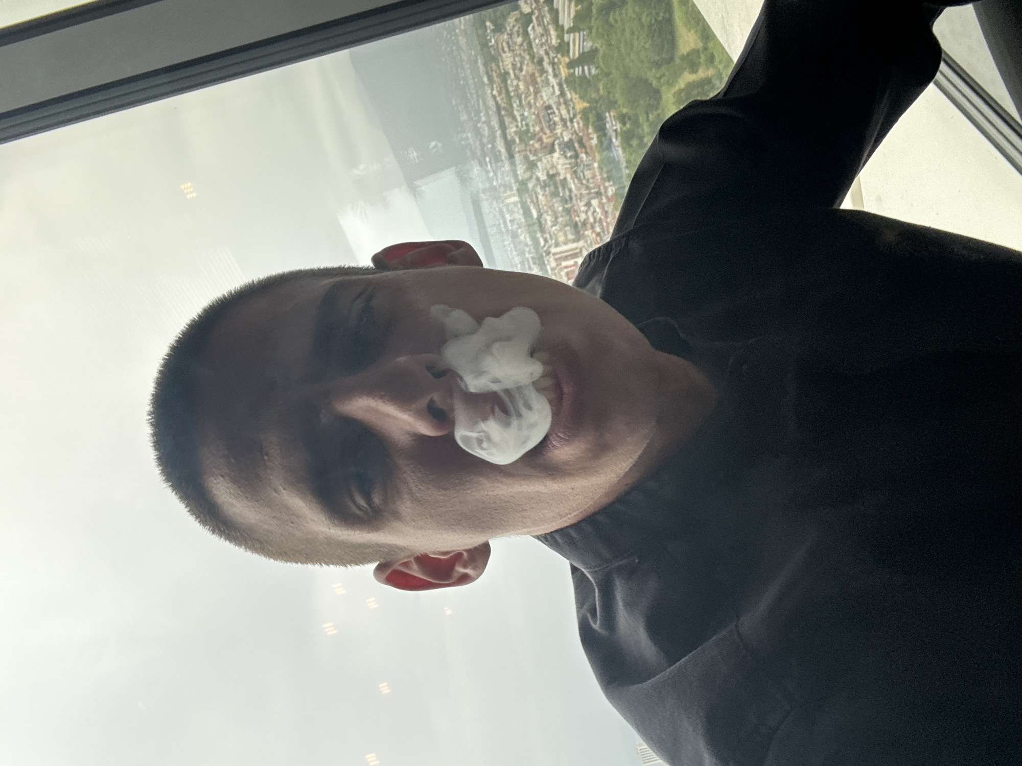 A close-up, low-angle selfie shows a young adult with a buzz cut exhaling a cloud of white vapor from their mouth, obscuring their lower face. They are wearing a black collared shirt. The person is positioned in the foreground, with a large window behind them. Through the window, a panoramic view of Sofia, Bulgaria, is visible, featuring a dense urban landscape with buildings, streets, and green trees. The sky is overcast, and there are some blurry reflections on the window. The image was taken indoors, likely on a high floor of a building overlooking the city.