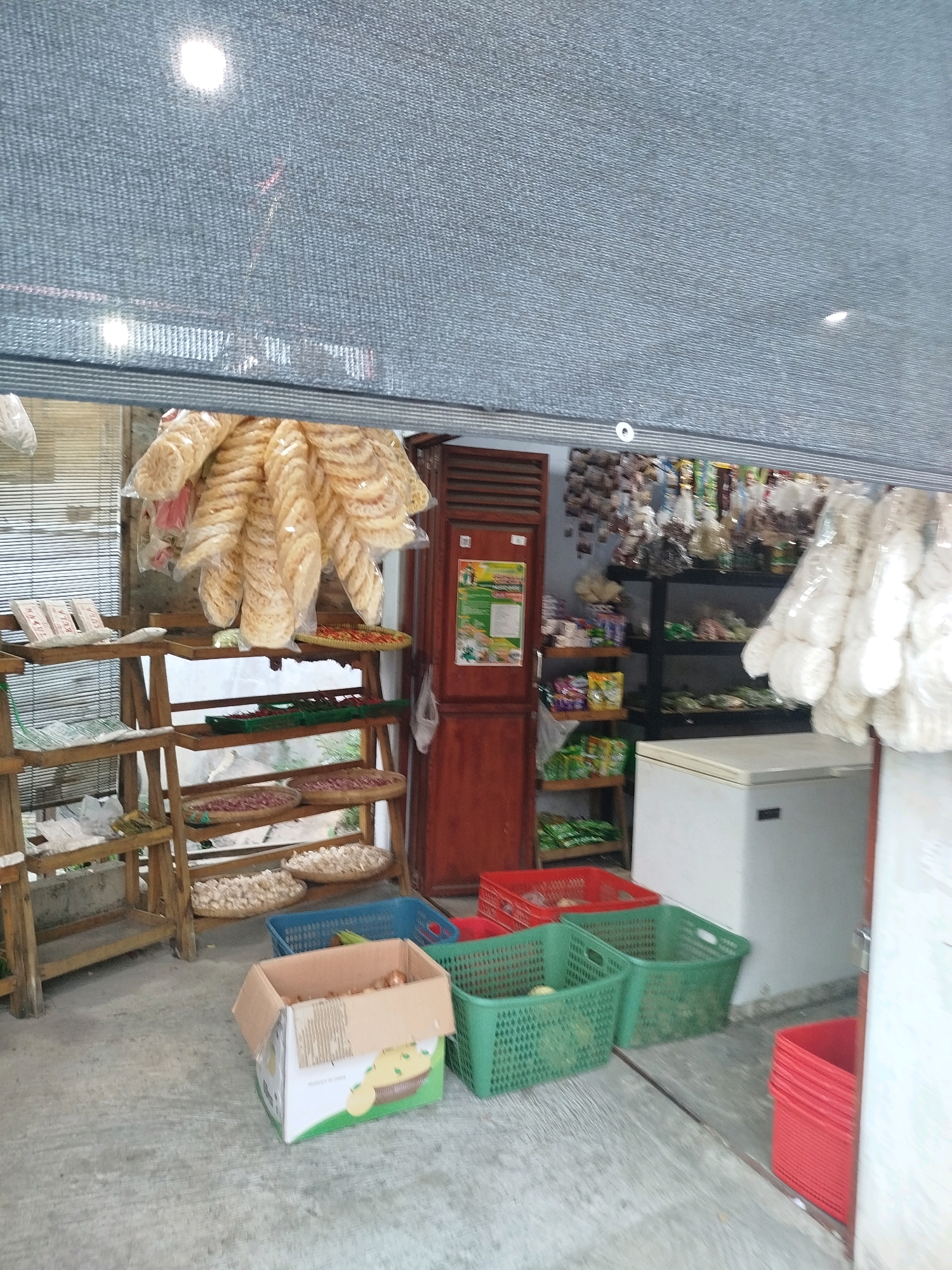 The image shows the interior of a small shop in Cekok, Indonesia. The shop appears to be a general store or a small grocery. In the foreground, there are several plastic baskets and a cardboard box filled with goods. Two green plastic baskets are in the center, one containing a large, round, green item, possibly a fruit or vegetable, and the other appearing to hold various food items. A red plastic basket is to the right, and another red basket is behind the green ones. A cardboard box, partially open, sits in the lower left, containing what appear to be onions or potatoes.
Behind the baskets, wooden shelves are stacked with products. The shelves hold various food items, including what appear to be dried goods, crackers, and possibly snacks. Some items are in plastic bags, while others are displayed loose in woven baskets. A large bunch of light-colored, circular crackers hangs prominently from the top shelf. A red-colored tray with some items on it is also visible on a shelf.
To the right of the shelves, a white chest freezer stands against the wall. Further back, more shelves are visible, stocked with packaged goods and more hanging items. A reddish-brown wooden door is on the far right. The walls appear to be a light color, and the floor is concrete. A gray mesh screen is draped across the top of the scene, obscuring the upper portion of the shop and letting in some light. The overall impression is of a modestly stocked, busy local shop. There are no people visible in the image.