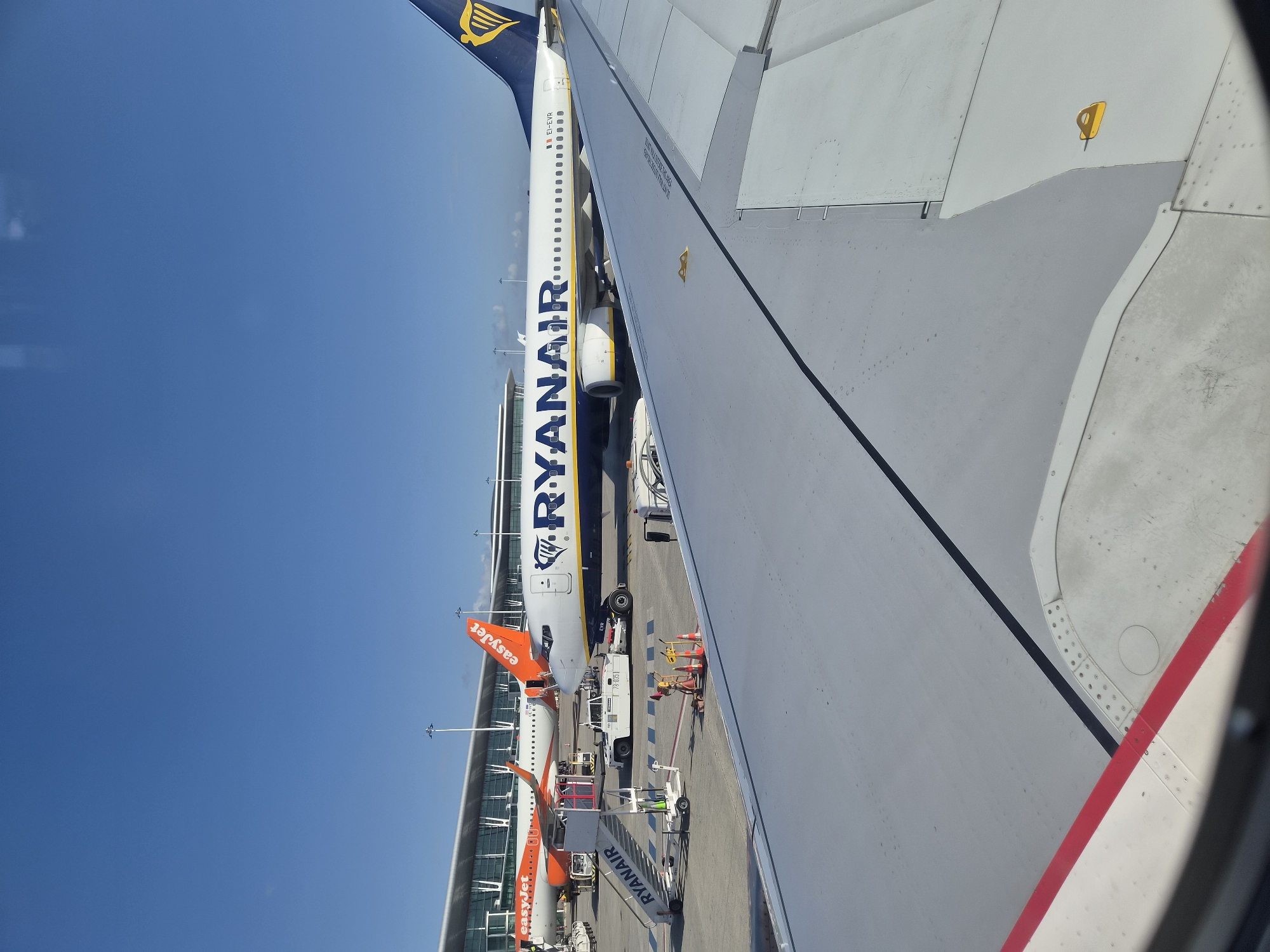 The image shows a view from an airplane window. The wing of the plane occupies the right side of the frame, with a visible red and white stripe along the edge. Beyond the wing, on the tarmac, there is another airplane painted in the colors of the airline "Ryanair" and an "EasyJet" plane behind it, along with ground support equipment, including stairs and vehicles. The terminal building is visible in the background. The sky is clear and blue. The location is in Moreira, Portugal, identified by the presence of Ryanair and EasyJet airlines, which are frequent at Portuguese airports.