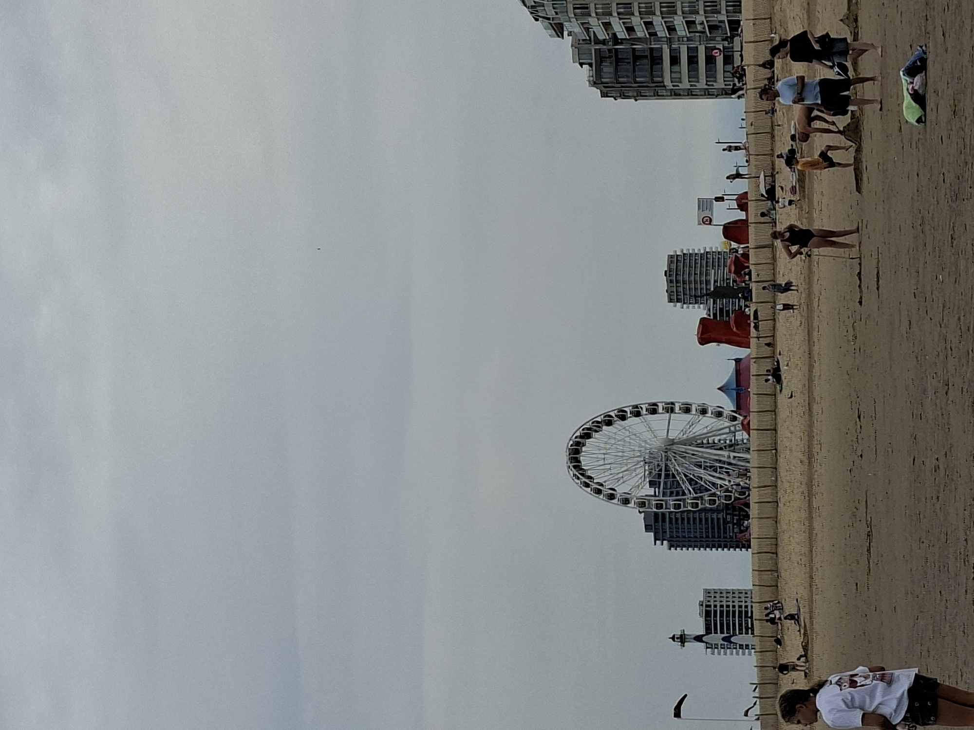 The image depicts a section of the beach in Ostend, Belgium, on an overcast day. In the foreground, several people are visible on the sandy beach. A young woman wearing a white t-shirt and dark shorts is seen in the left of the frame, facing away from the camera. Further into the scene, a group of individuals are engaged in various activities, including building sandcastles and standing around. A woman in a black swimsuit is near a shovel, and a man is helping a boy with sand. Another couple stands together further to the right.
In the mid-ground, a wooden fence separates the beach from a promenade or park area. Behind the fence, there are several structures. A large Ferris wheel dominates the skyline, with a lighthouse-like building to its left. Further to the right, several modern apartment buildings with balconies are visible, characteristic of the Ostend coastline. Two large, abstract red sculptures are also present in this area. The sky is uniformly gray and cloudy, suggesting a temperate climate. The overall scene captures a moment of leisure activity on a beach with urban elements in the background.